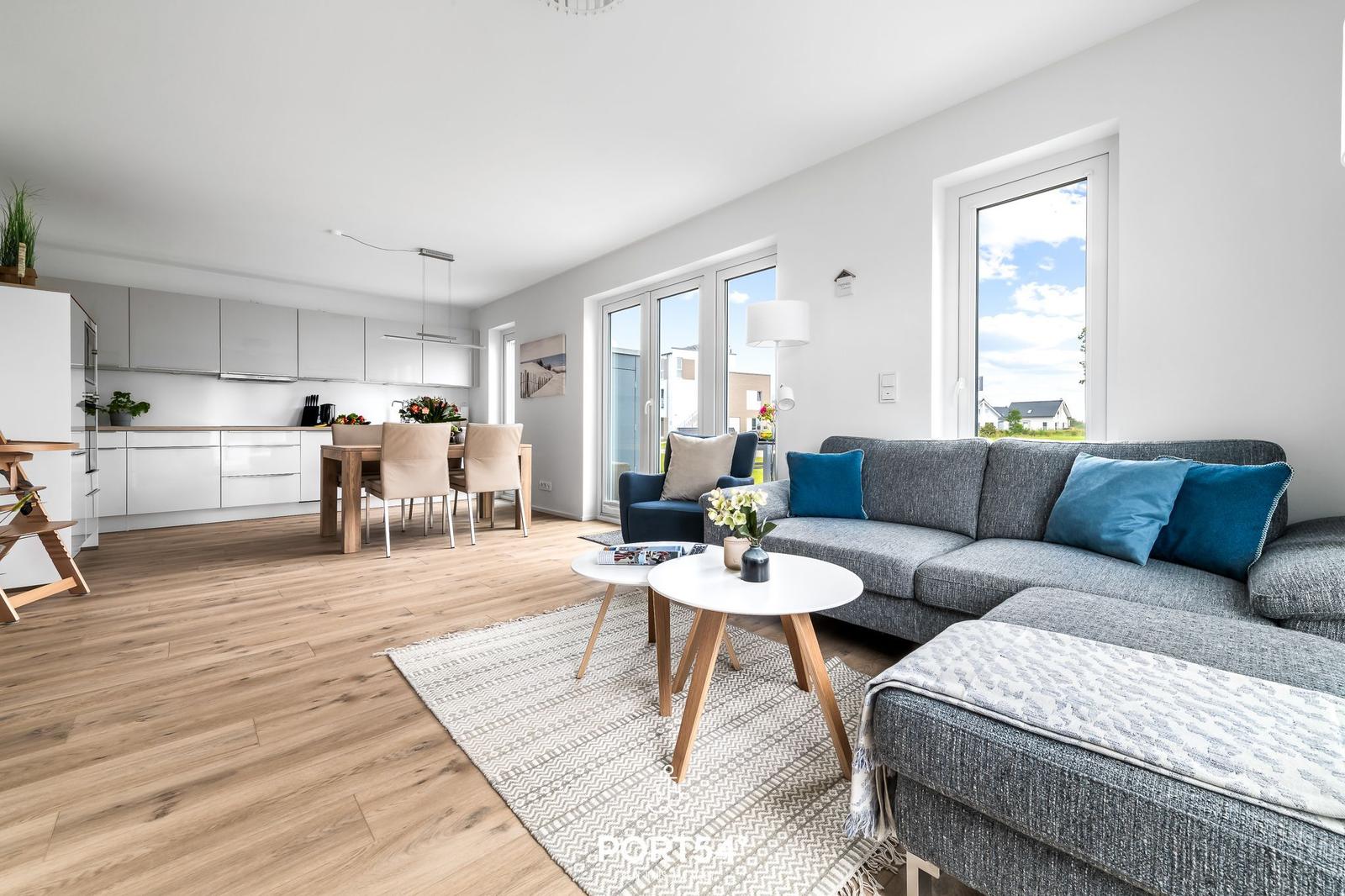 Wohn- und Essbereich mit Küchenzeile und großem Fenster. Holzboden und graue Couch mit blauen Kissen.