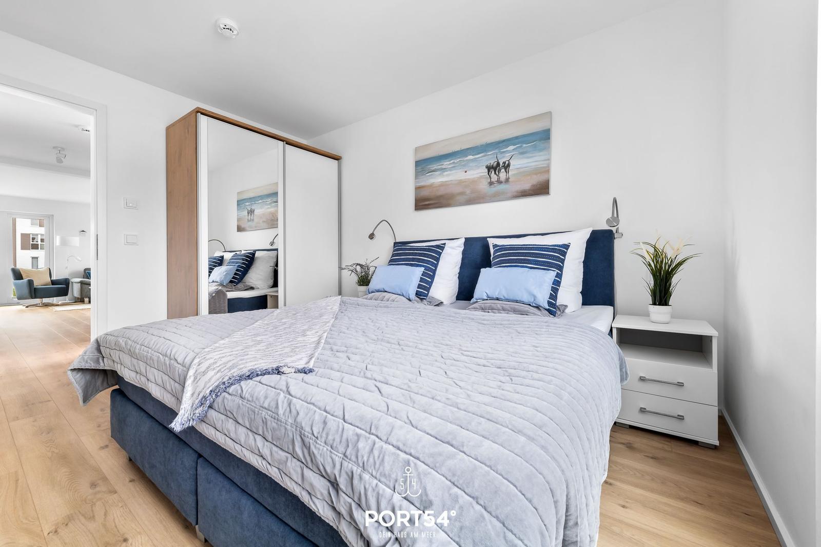 Schlafzimmer mit Bett, Nachttisch und Kleiderschrank. Wandbild und Holzboden sichtbar.