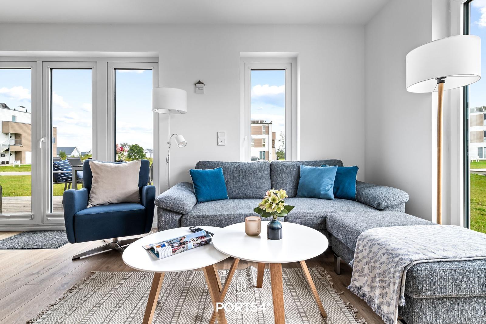 Wohnzimmer mit grauem Sofa, blauen Kissen und zwei weißen Tischen. Große Fenster und Terrasse.