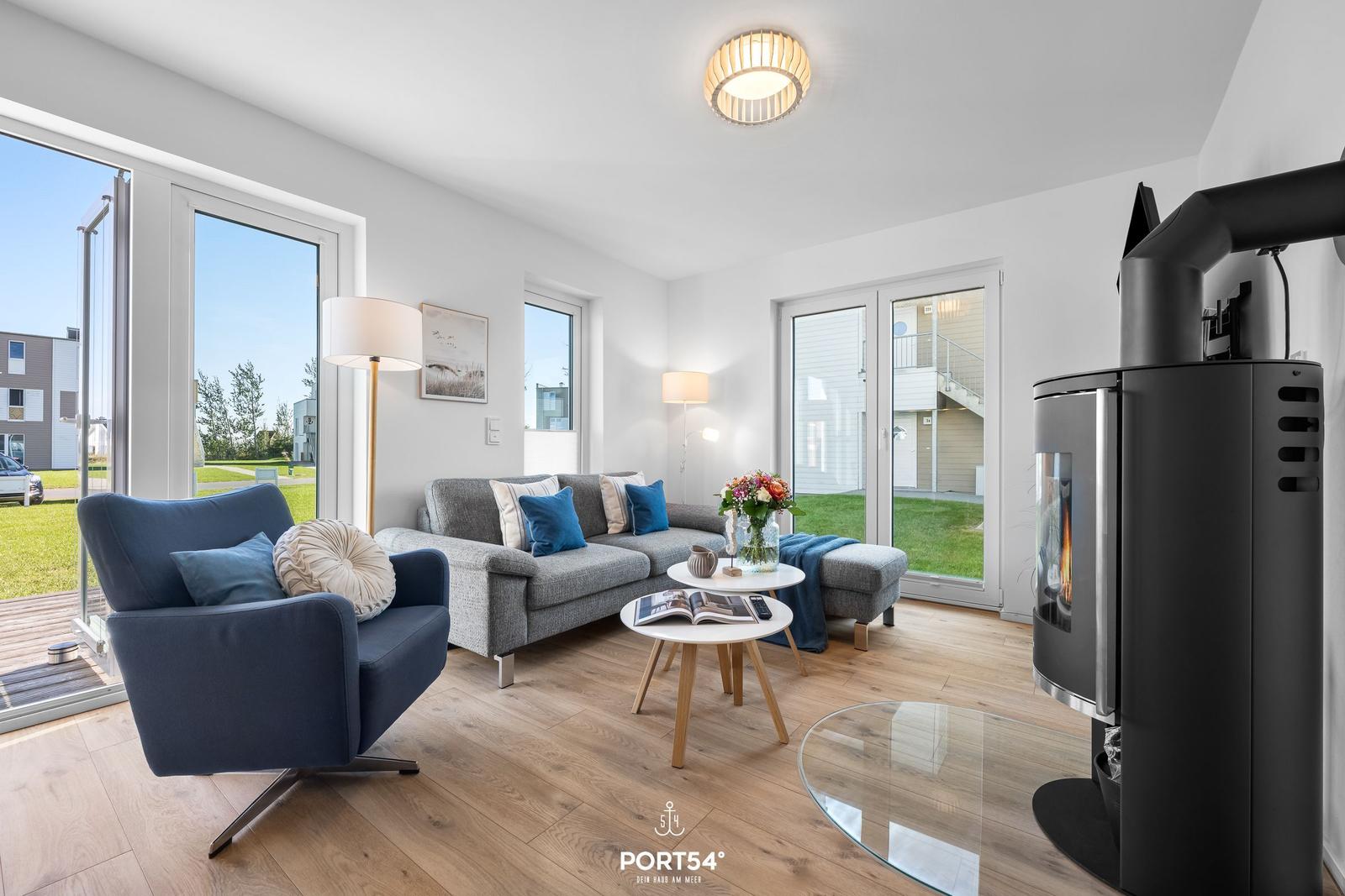 Wohnzimmer mit Sofa, Sessel und Kaminofen. Große Fenster und Holzboden.