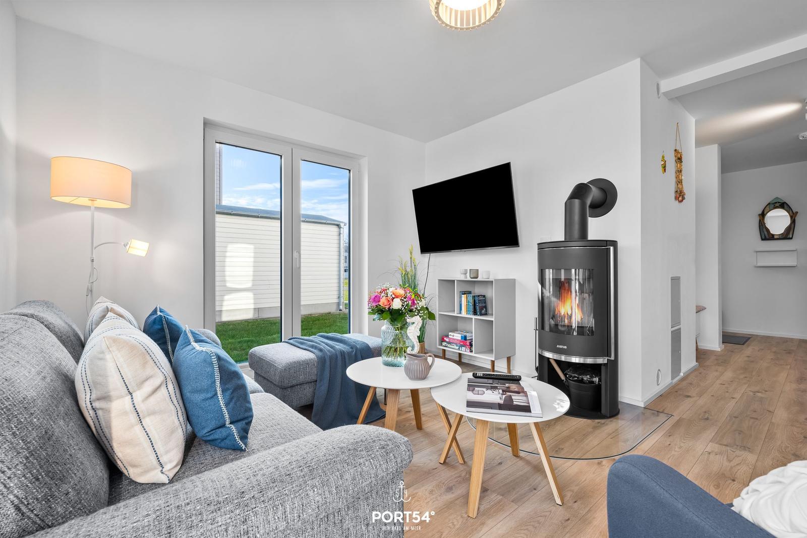 Wohnzimmer mit Sofa, Kaminofen, Fernseher und Glasfront zur Terrasse.