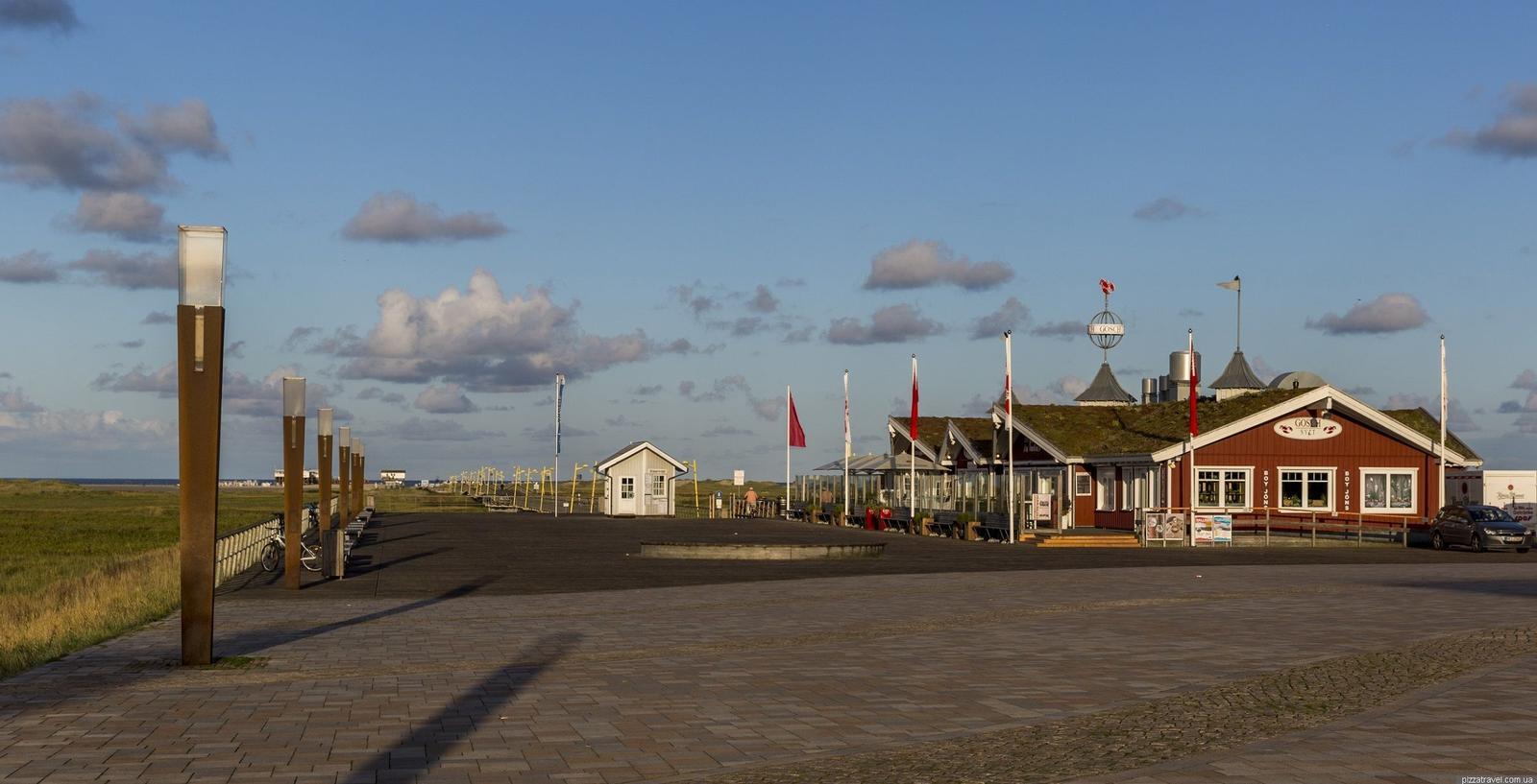 Pfad mit Laternen führt zu Gebäuden mit Grasdächern und Flaggen unter blauem Himmel.