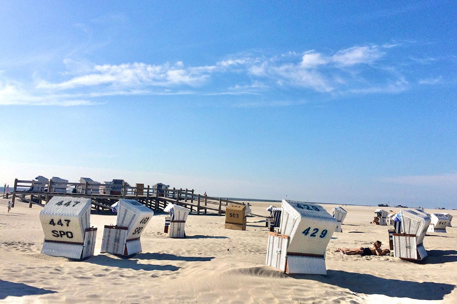 Strand mit weißen Strandkörben und Holzsteg unter blauem Himmel.