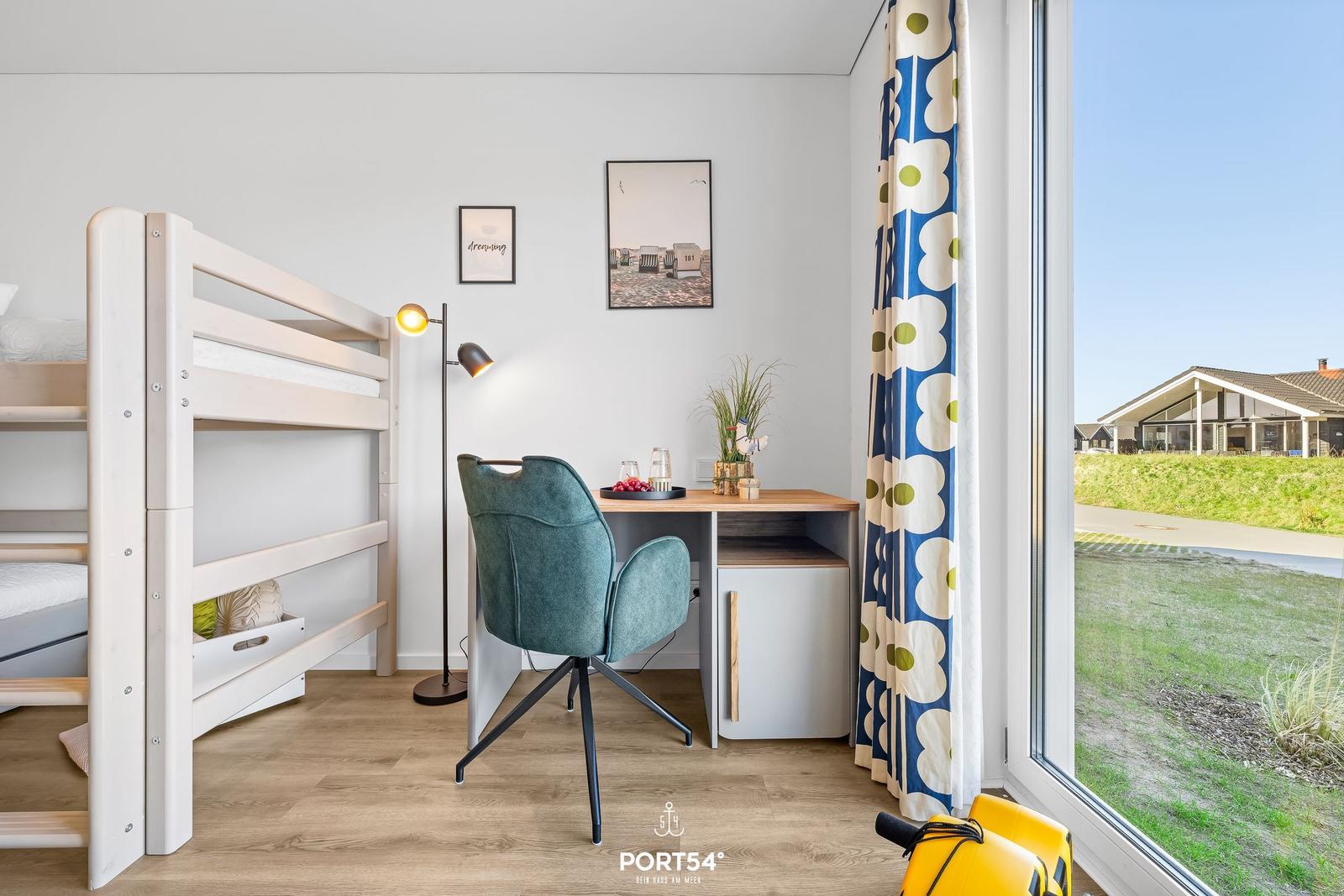 Ein Raum mit Doppelstockbett, Schreibtisch und Stuhl. Durch ein Fenster sieht man eine grüne Landschaft.