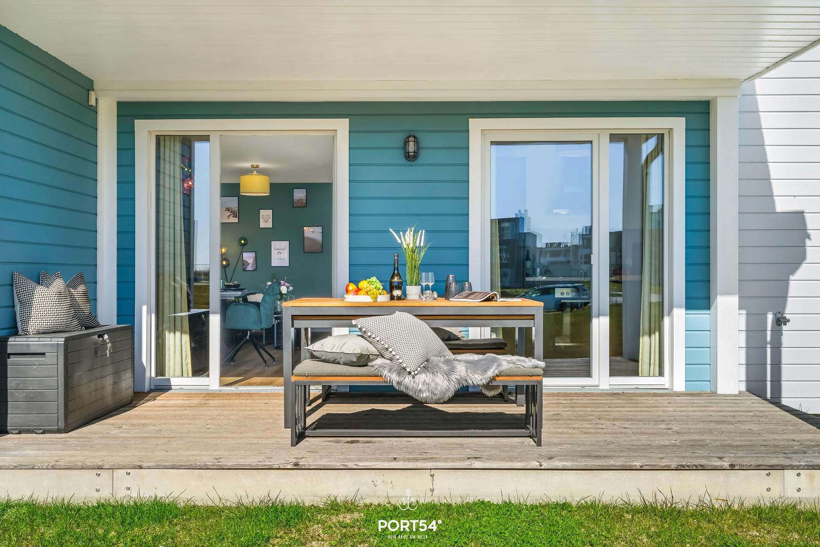 Terrasse mit Tisch, Bank und Blick ins Haus. Holzdeck und blaue Fassade.