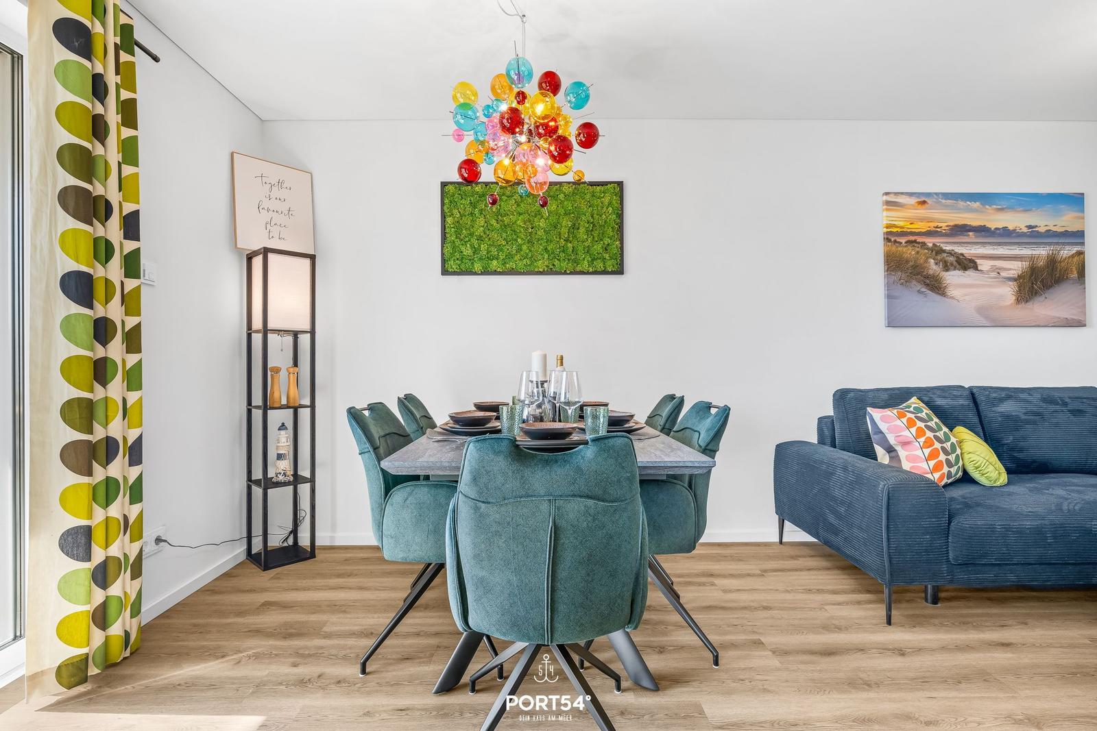 Esszimmer mit Esstisch, sechs Stühlen und Sofa. Deckenleuchte und Wandbilder sichtbar.