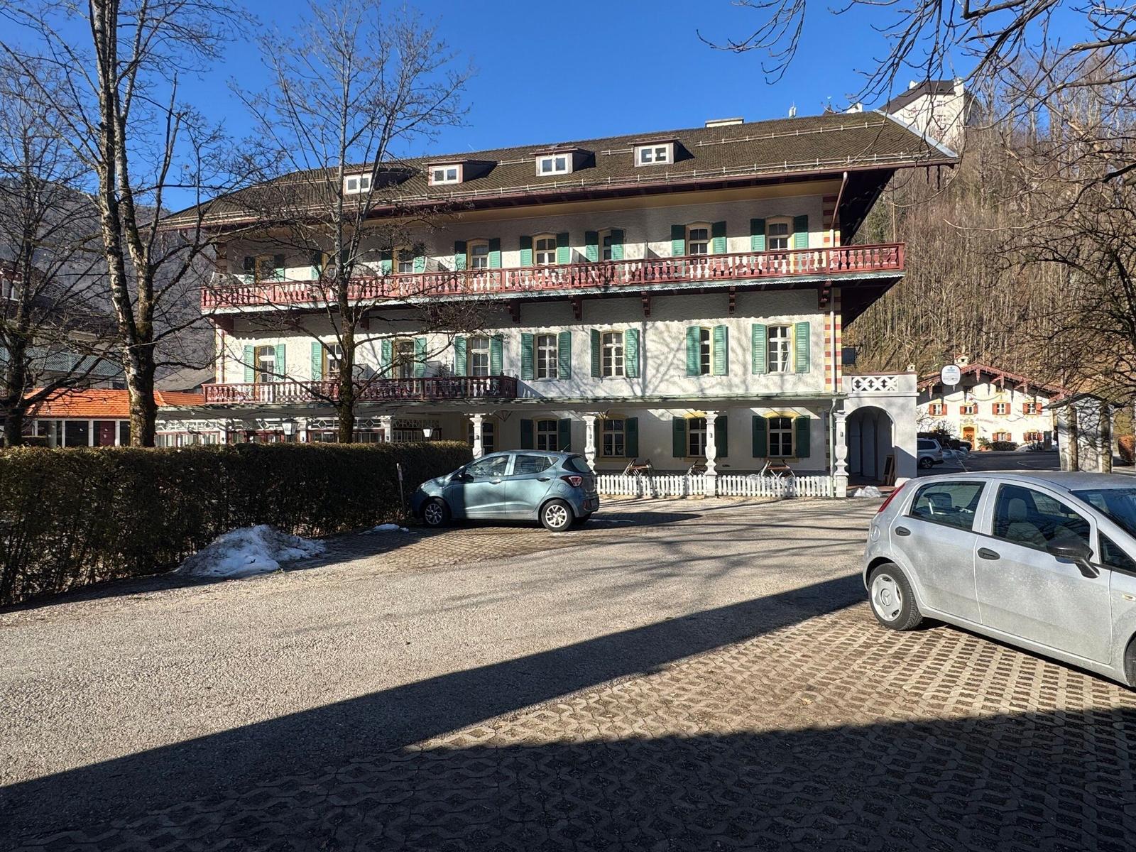 Ein großes Haus mit zwei Balkonen und grünen Fensterläden. Zwei Autos stehen auf dem Hof.