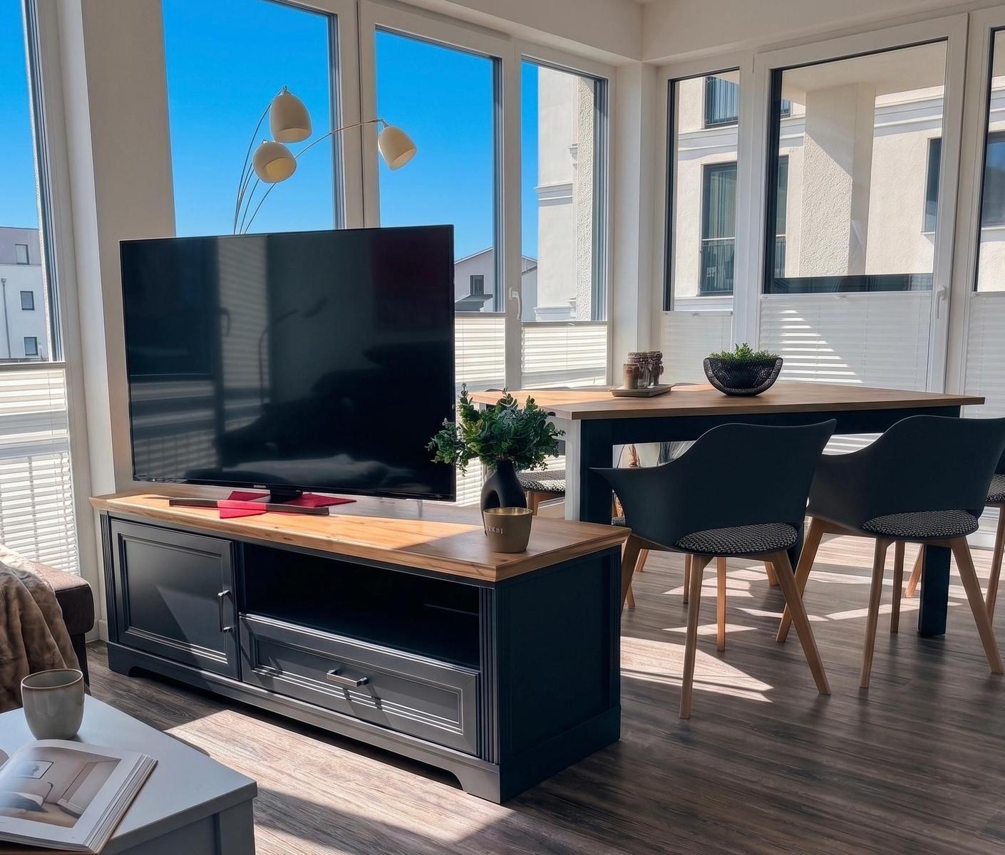 Wohn- und Essbereich mit TV, Tisch und Stühlen. Große Fenster mit Blick auf Gebäude.
