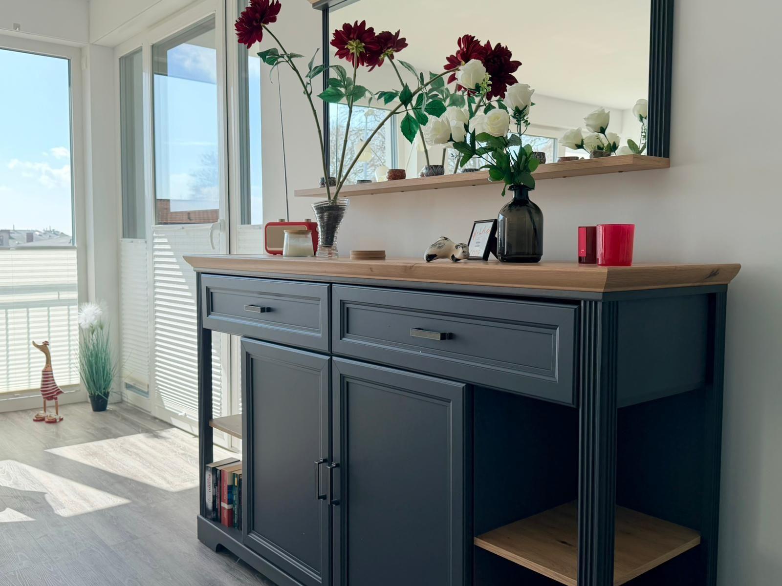 Schwarzer Sideboard mit Holzoberfläche, Blumen und Dekoration. Große Fenster mit Blick auf Balkon.