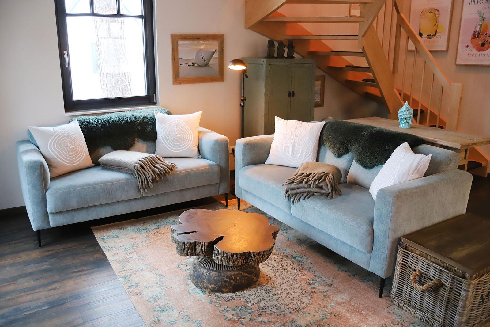 Zwei graue Sofas mit Kissen und Decken stehen auf einem Teppich. Eine Holztreppe führt nach oben.