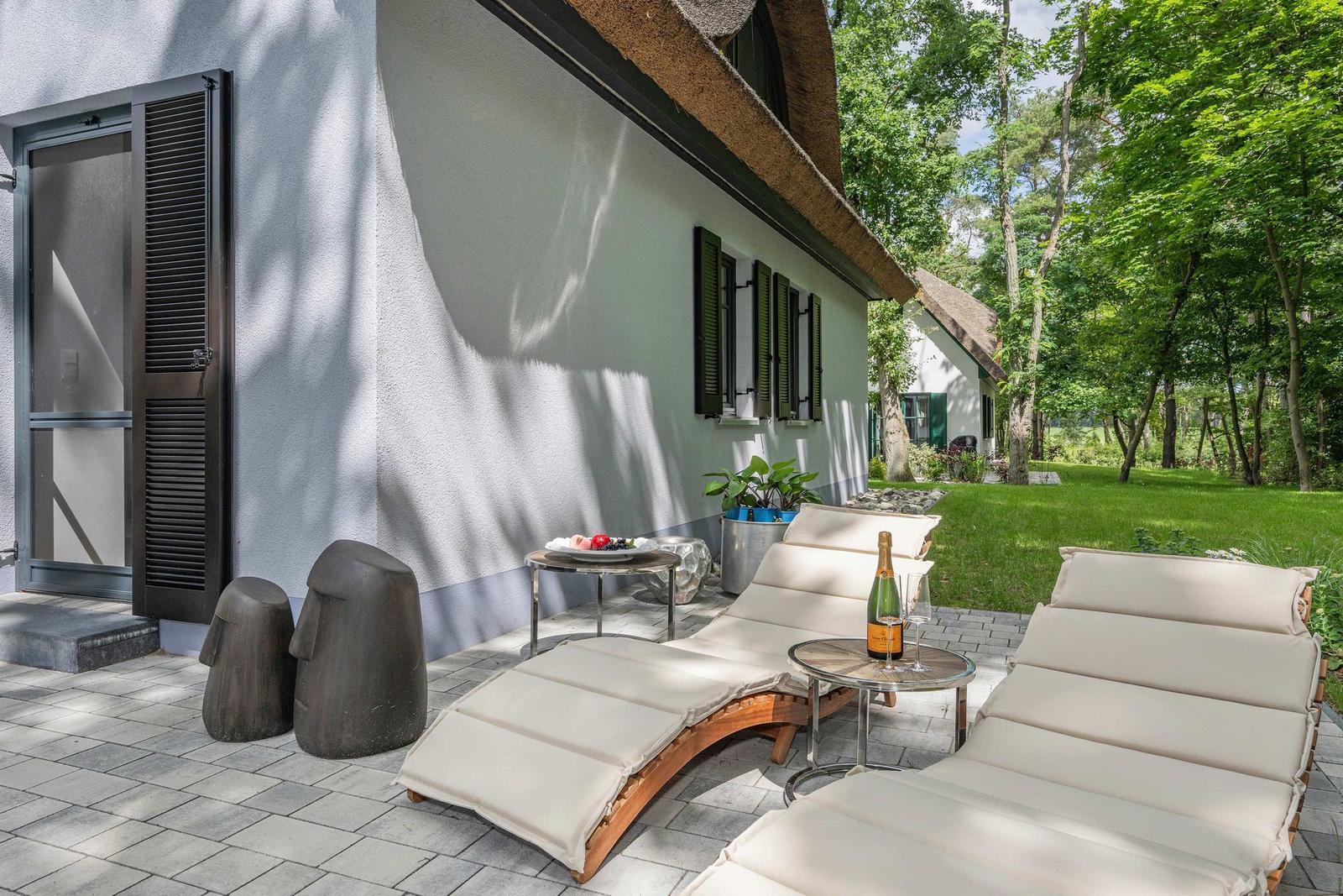 Terrasse mit Liegen, Tisch und Flasche. Hinterhof mit Grünfläche und Bäumen.