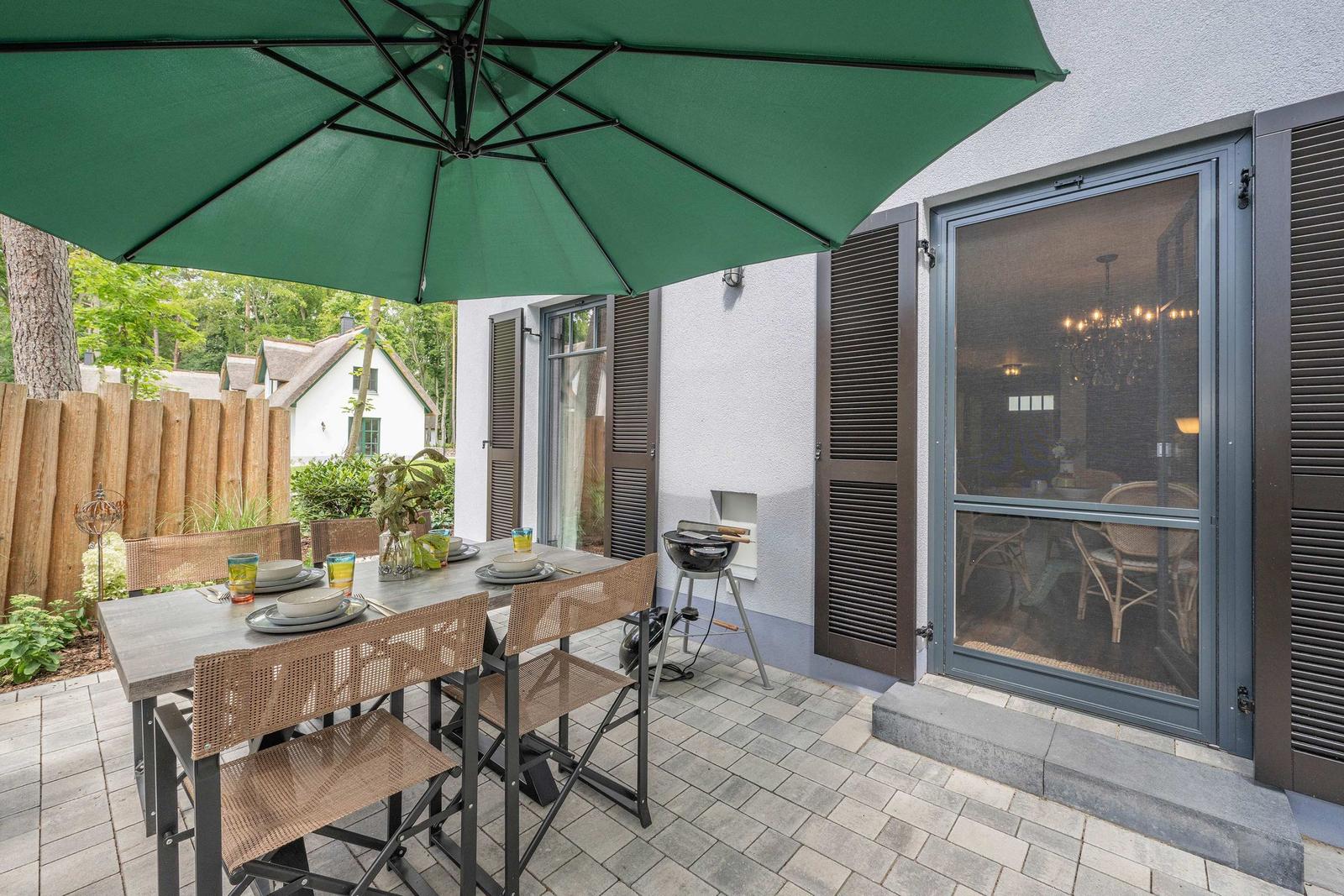 Terrasse mit Tisch, Stühlen und grünem Sonnenschirm. Grill und Glastür zur Inneneinrichtung.