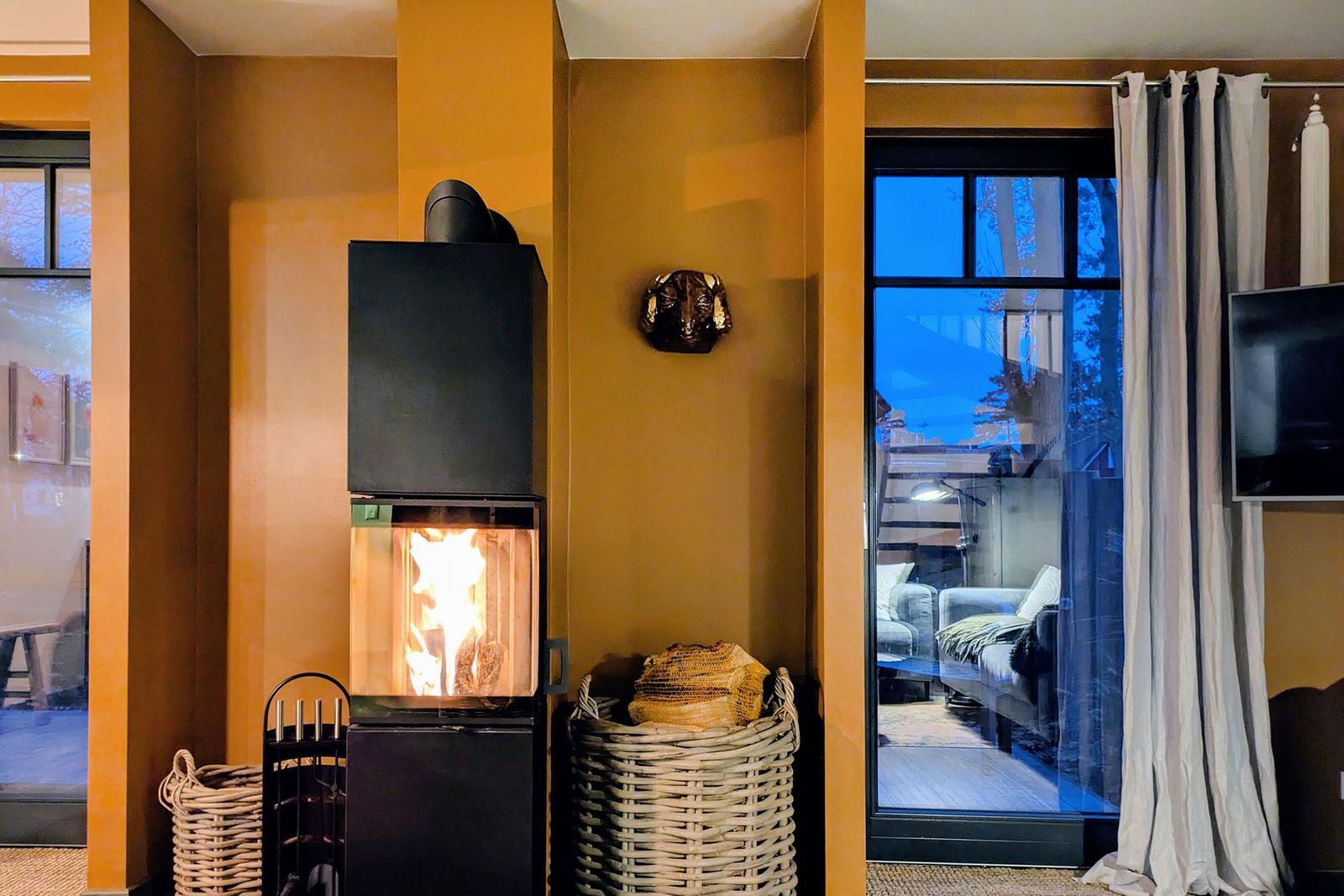 Ein Kamin mit brennendem Holz, Körbe und eine Glasfront zur Terrasse.