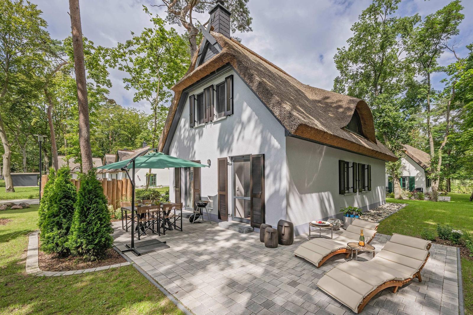 Haus mit Strohdach, Terrasse mit Liegen und Tisch, umgeben von Bäumen.
