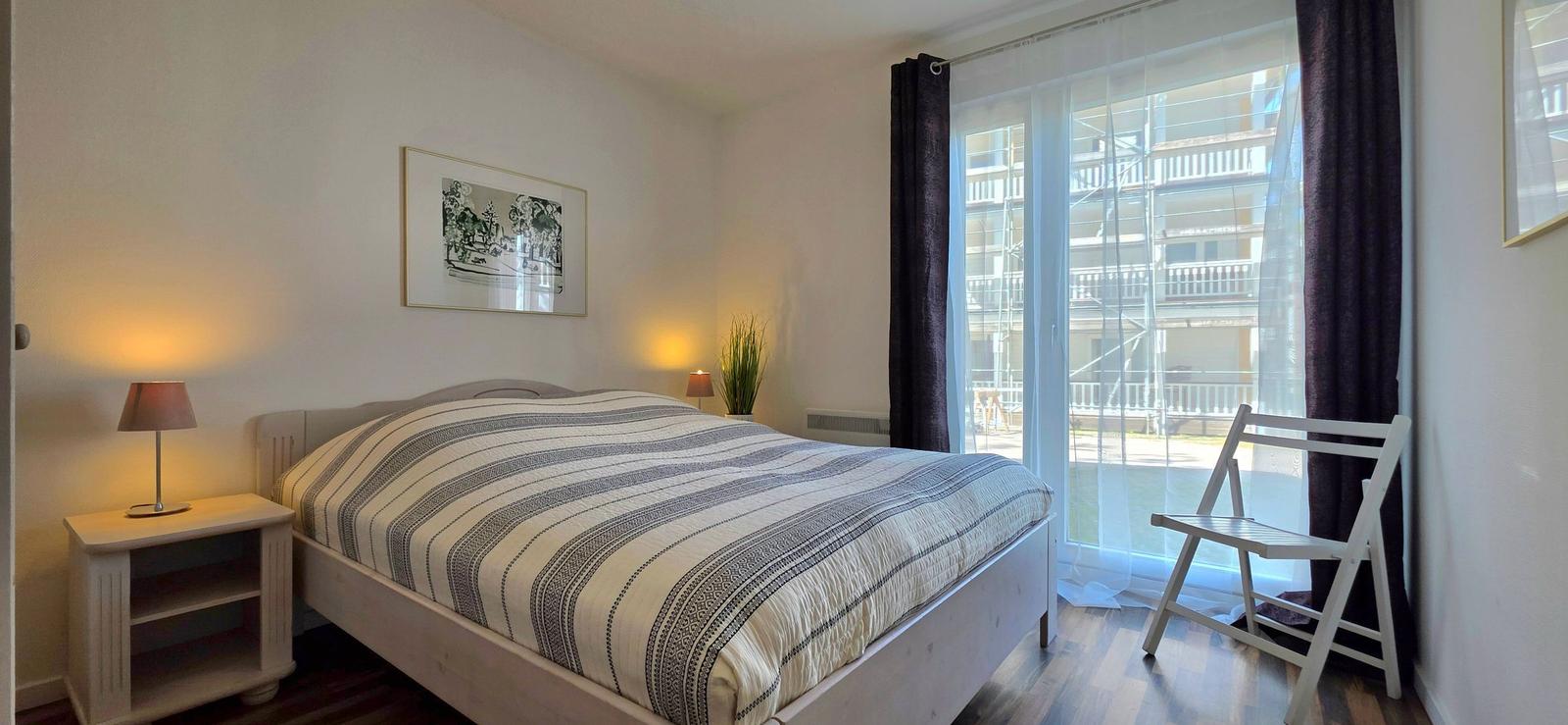 Schlafzimmer mit Bett, Nachttisch und Stuhl. Durchgangstür führt auf Balkon.
