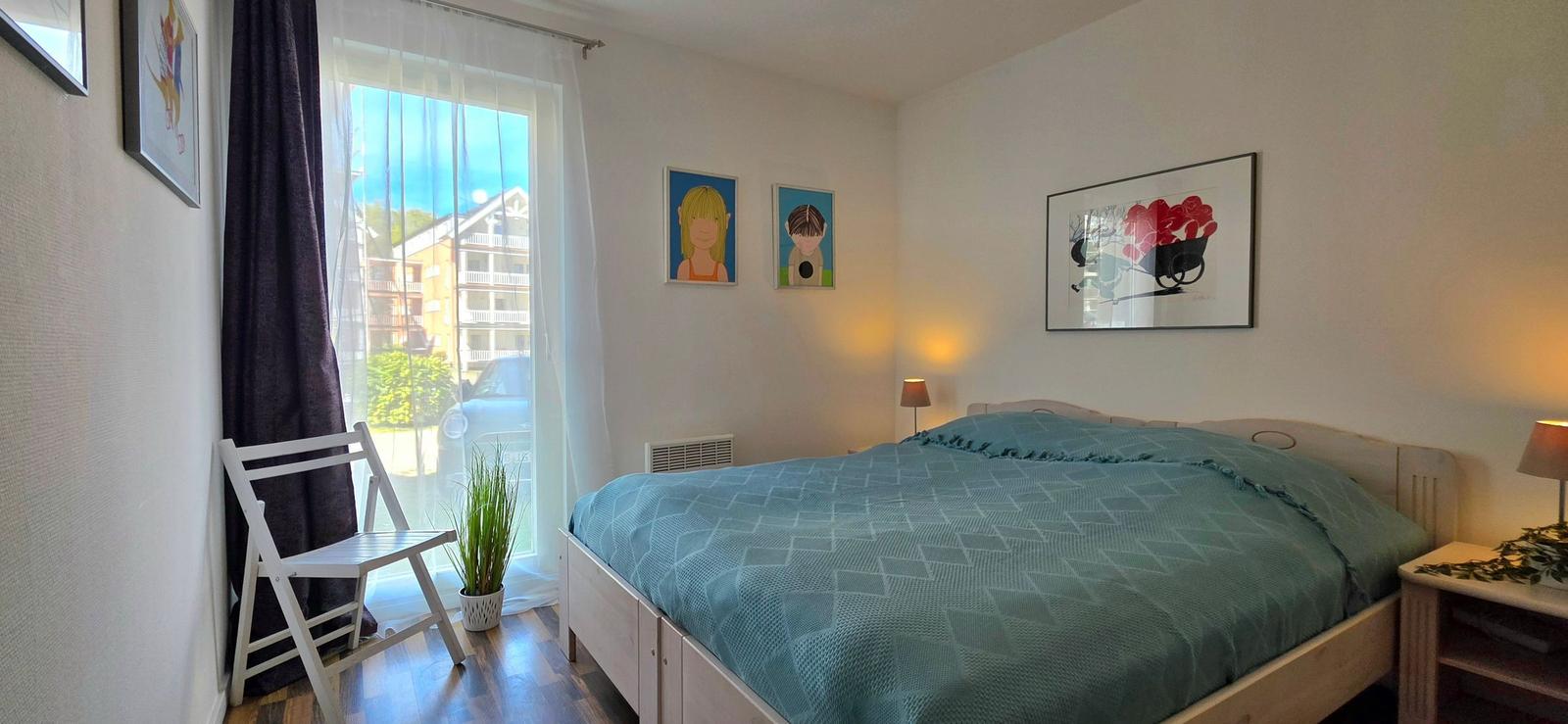 Schlafzimmer mit Bett, Fenster, Stuhl und Wandbildern.