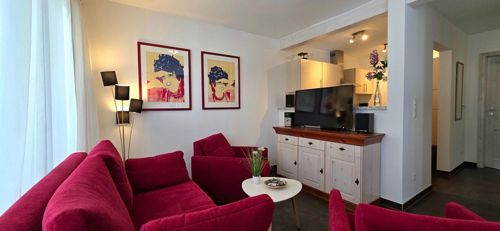 Wohnzimmer mit rotem Sofa, Fernseher und Küchenbereich im Hintergrund.