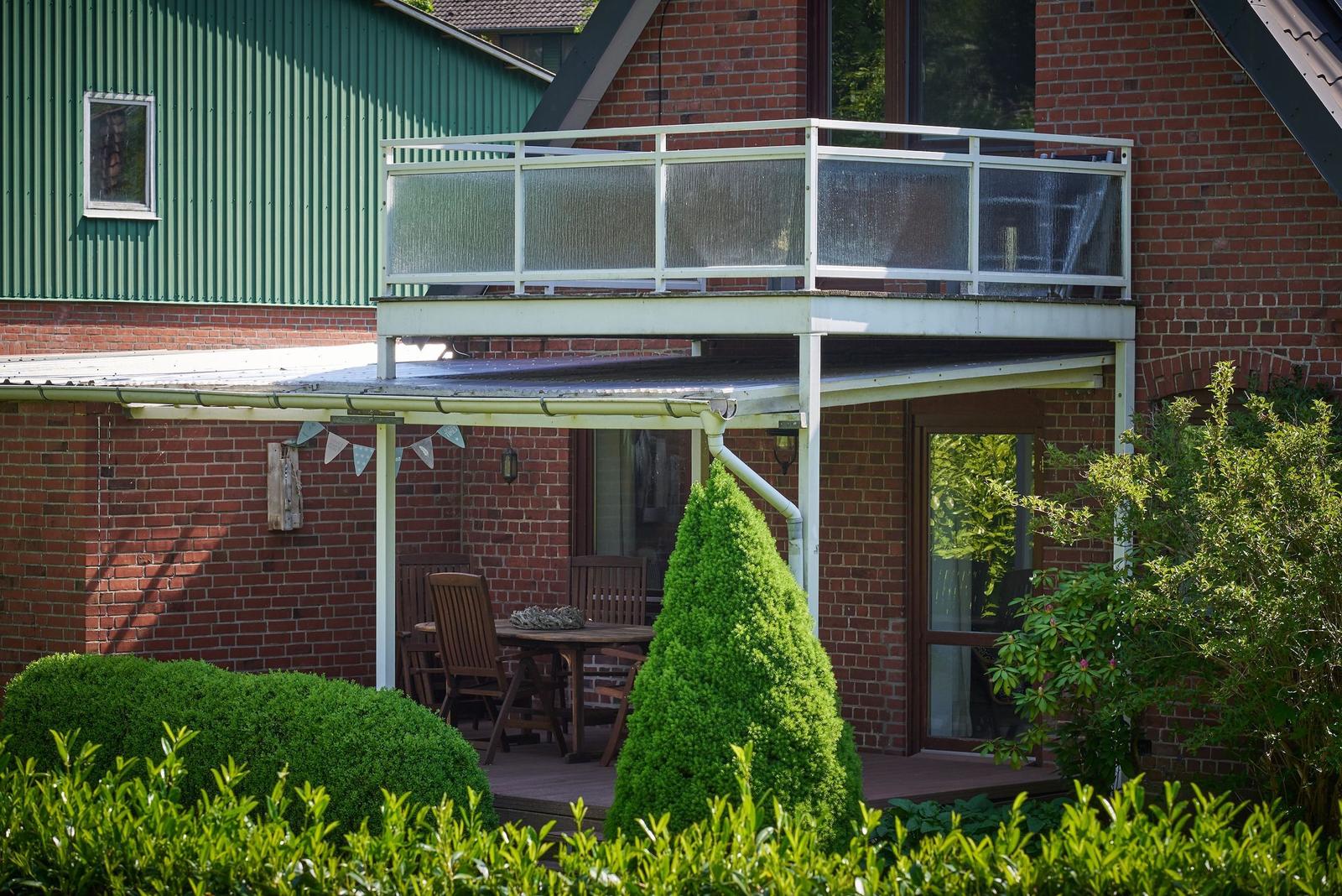 Terrasse mit Tisch und Stühlen, umgeben von grünen Sträuchern und einem Holzzaun.