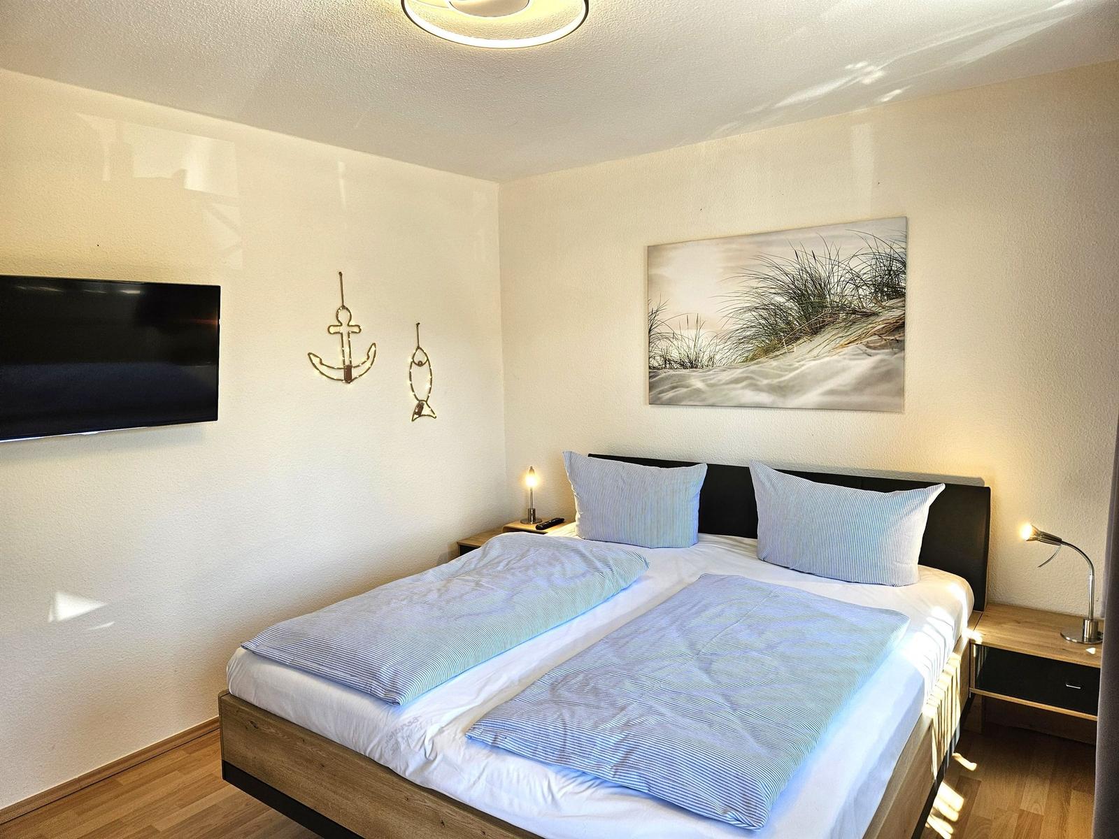 Schlafzimmer mit Bett, Nachttischen und Wandbild. An der Wand hängt ein Fernseher und zwei Dekorationen.
