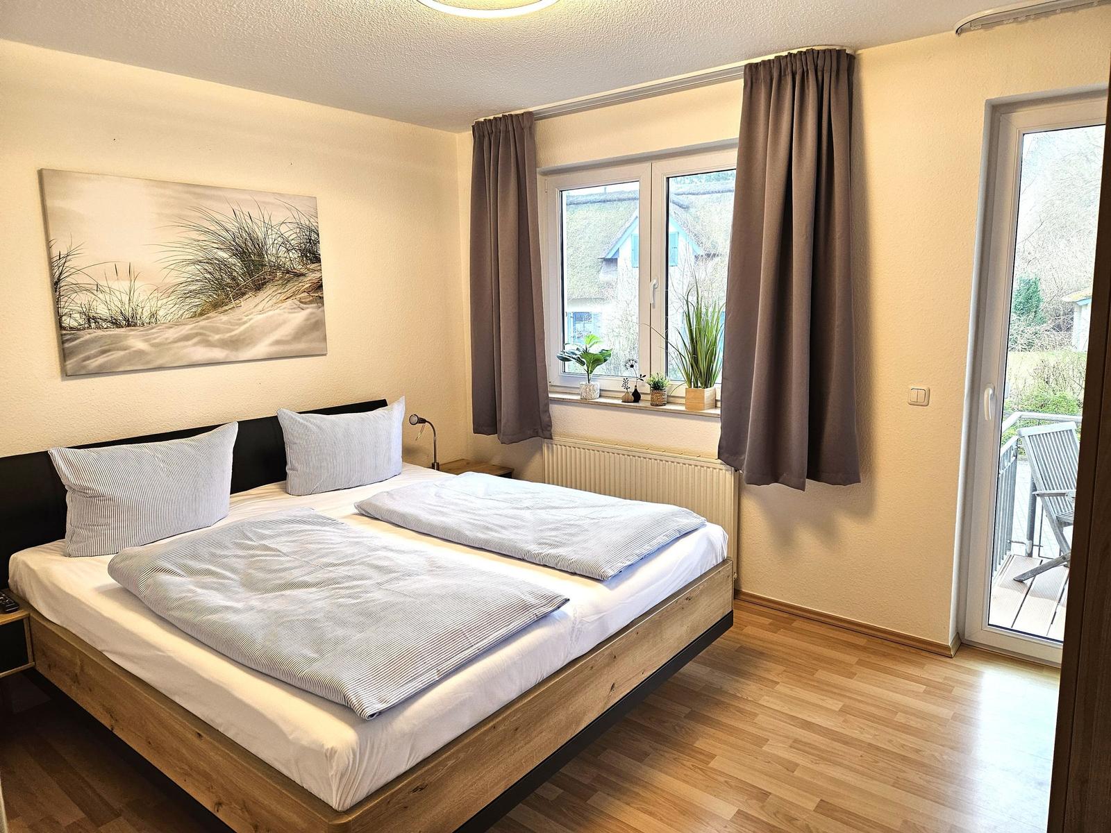 Schlafzimmer mit Doppelbett, Fenster und Balkontür. Wandbild und Pflanzen auf dem Fensterbrett.
