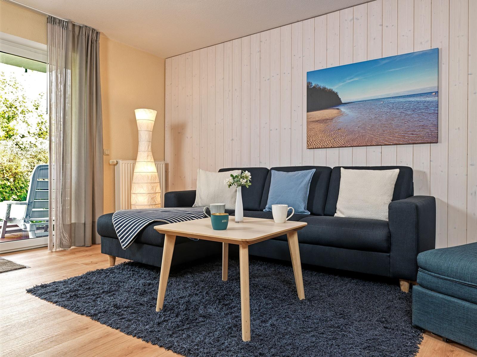 Ein Wohnzimmer mit Sofa, Tisch und Wandbild. Durchgang zur Terrasse.