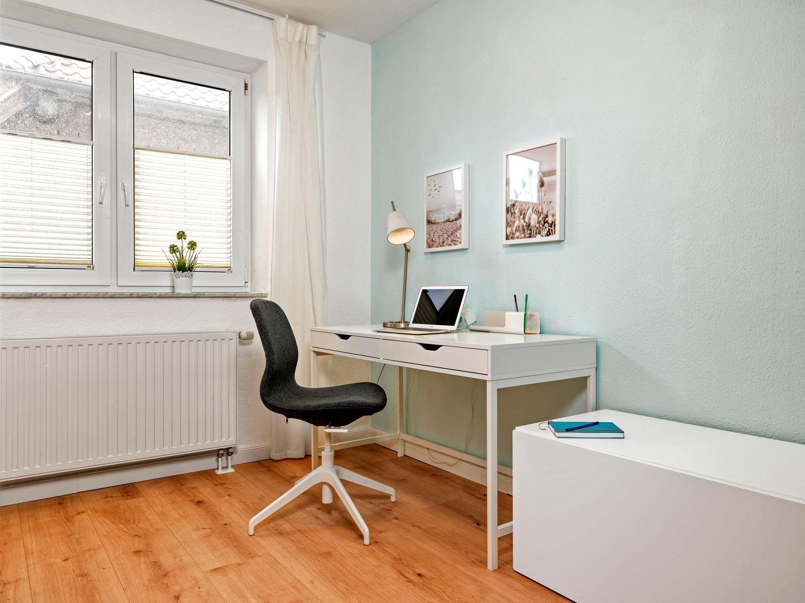 Ein Arbeitszimmer mit Schreibtisch, Stuhl und Fenster. Wandfarbe ist hellblau.