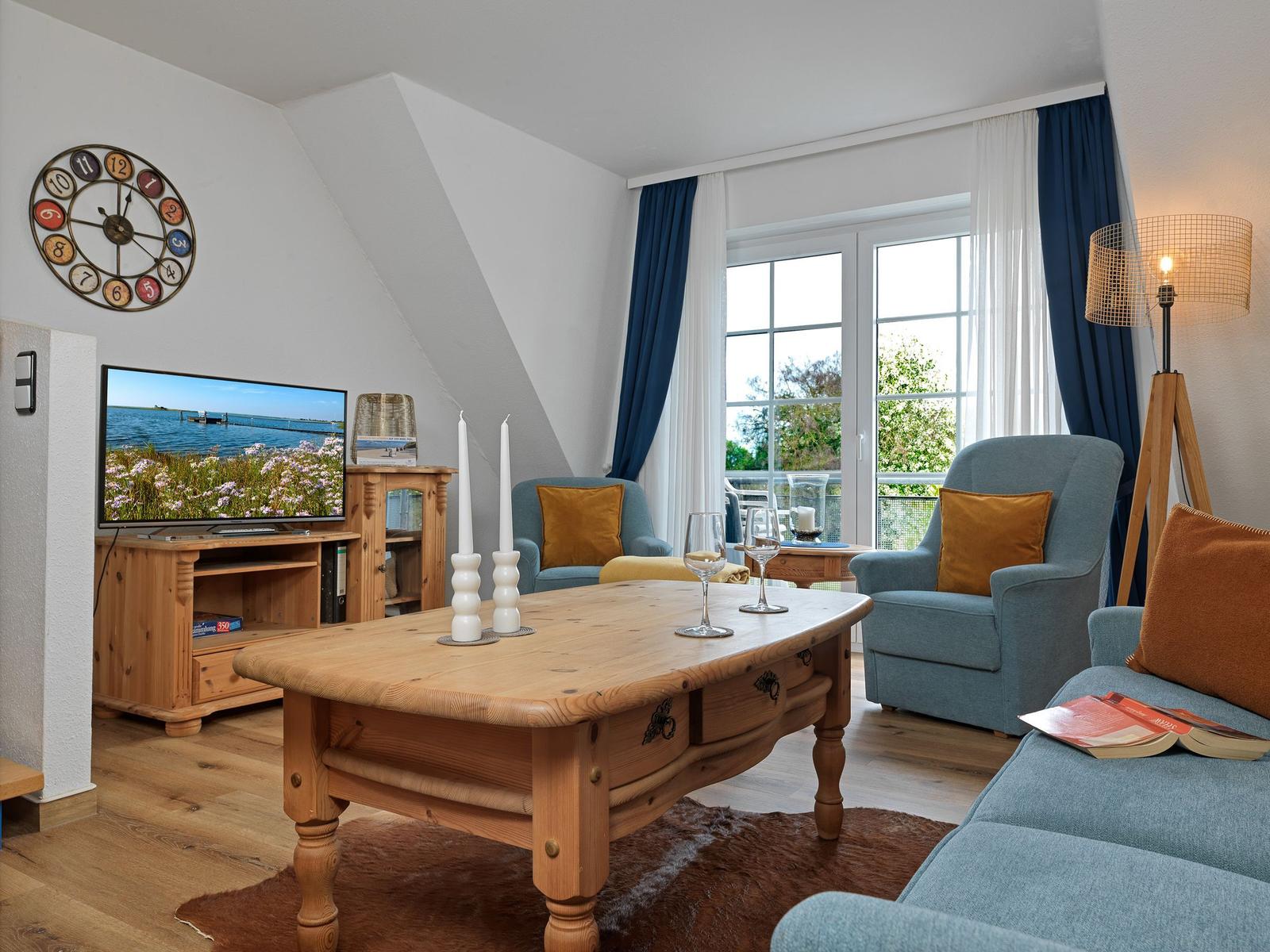 Wohnzimmer mit Holztisch, blauen Sesseln und TV. Fenster mit Blick auf Balkon.