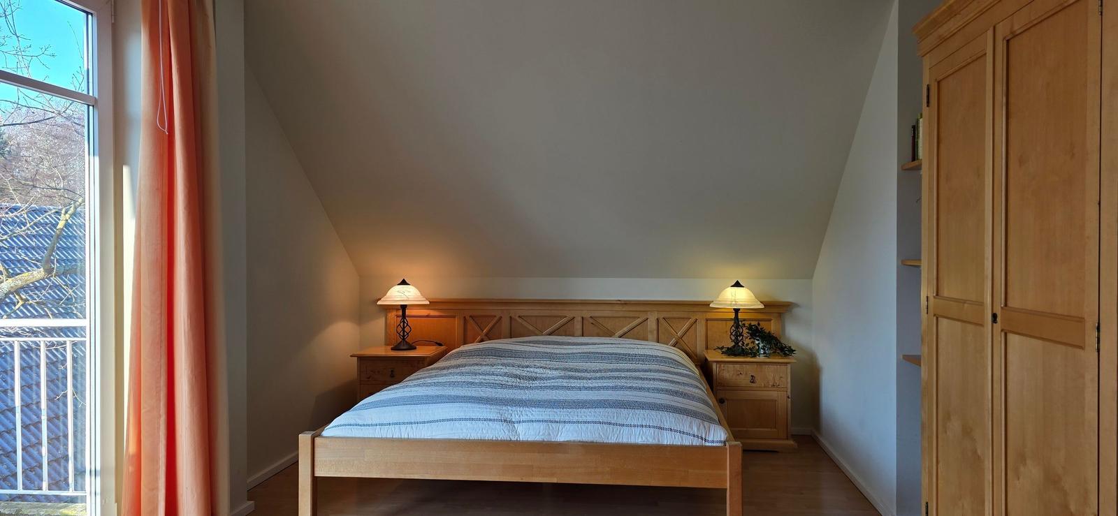 Schlafzimmer mit Holzbett, zwei Nachttischen und Schrank. Fenster mit orangefarbenen Vorhängen.