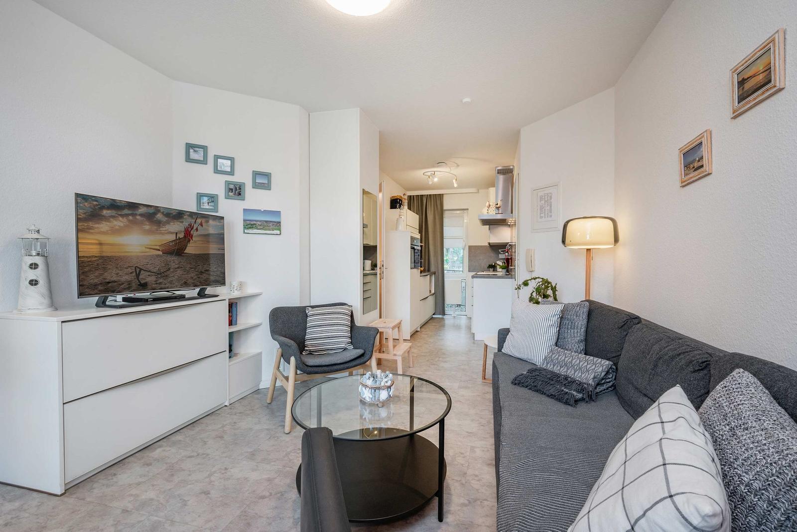 Wohnzimmer mit Sofa, Sessel und Glas-Tisch. Fernseher auf weißen Schrank. Küche im Hintergrund.