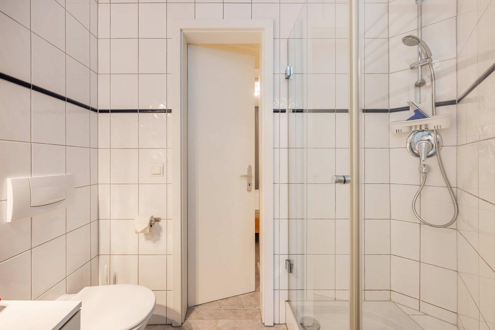 Badezimmer mit Dusche, Toilette und Tür zum angrenzenden Raum.