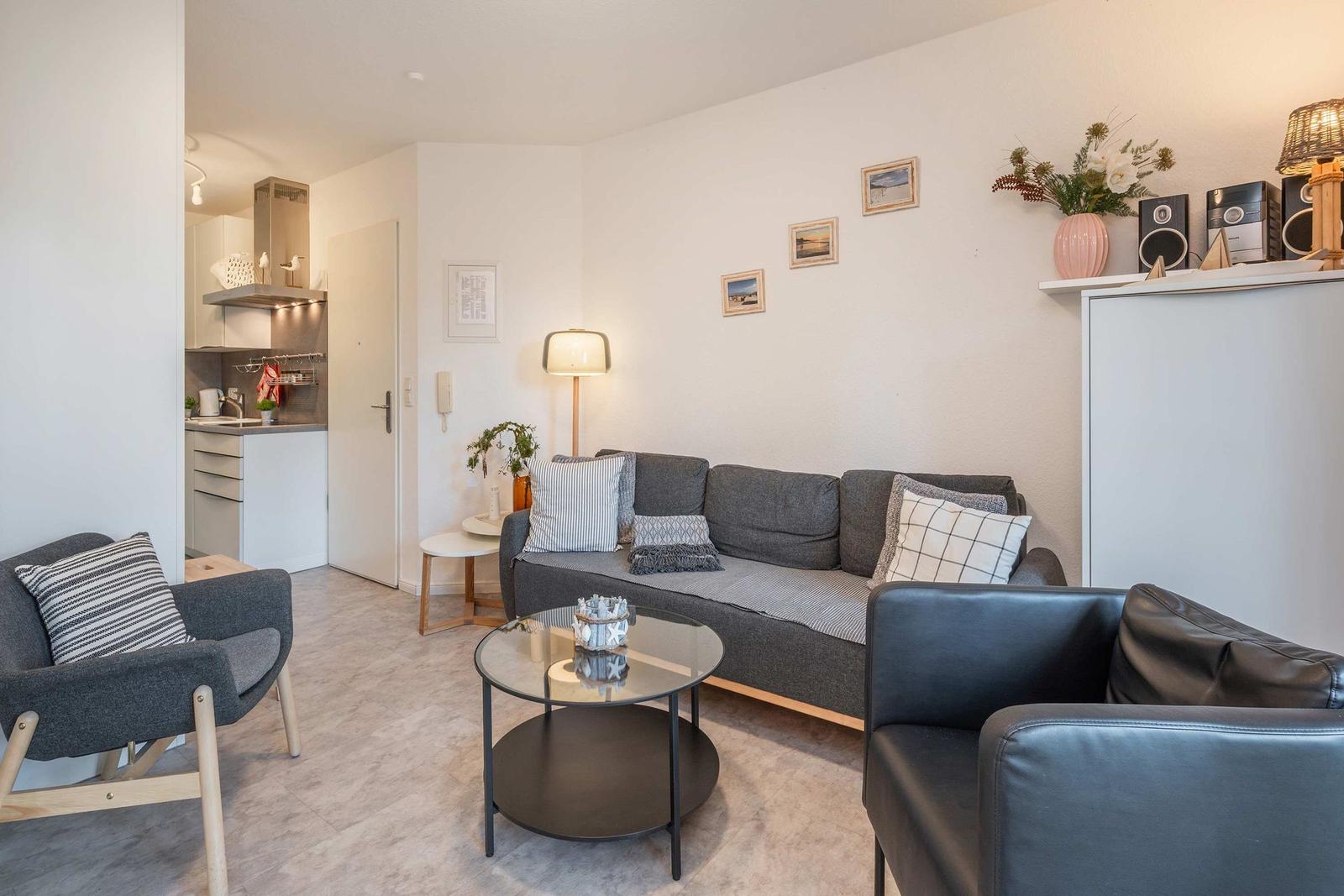 Wohnzimmer mit Sofa, Sessel und Couchtisch. Küche im Hintergrund.