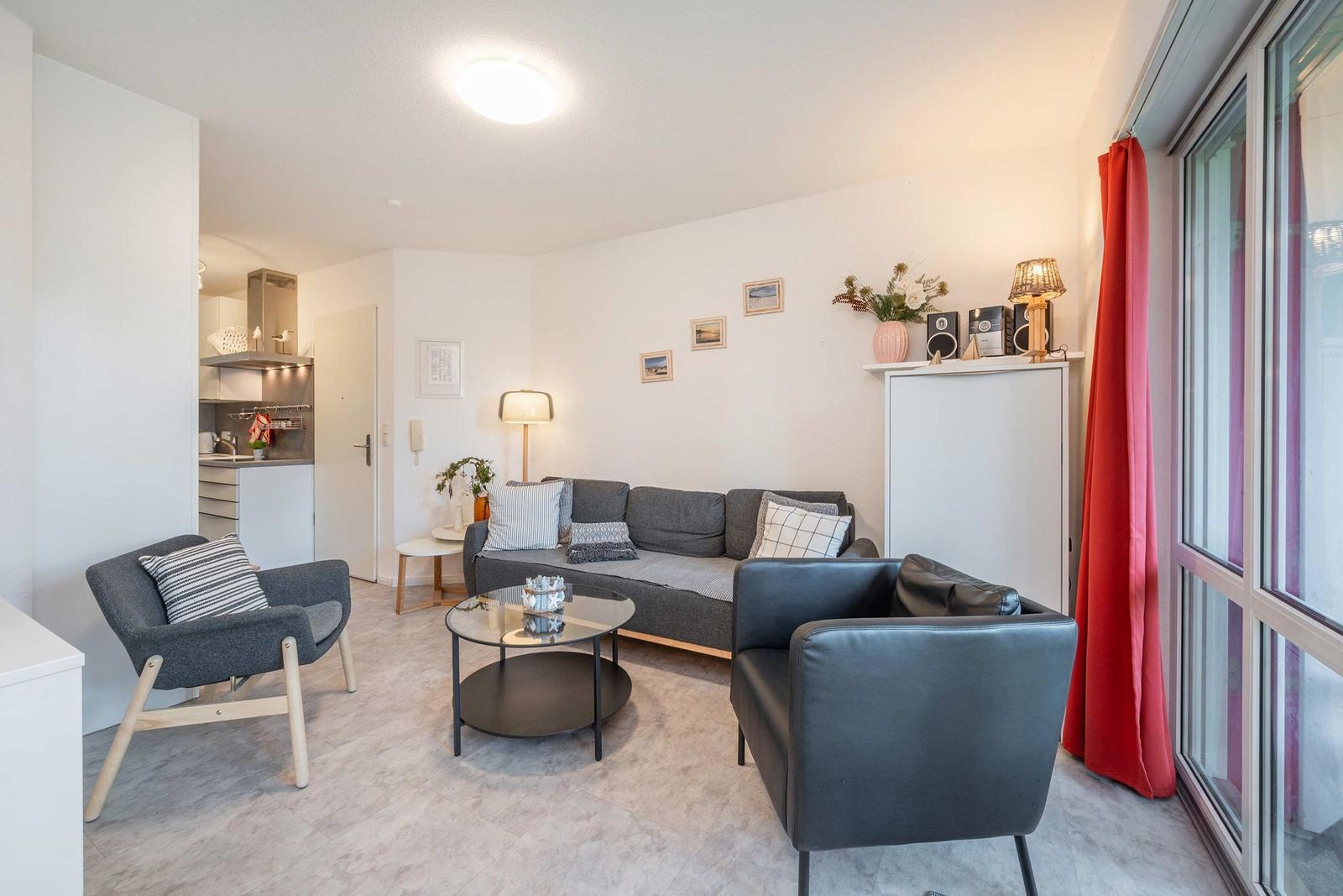 Wohnbereich mit Sofa, zwei Sesseln und Glas-Tisch. Küche im Hintergrund. Rote Vorhänge an Fenster.