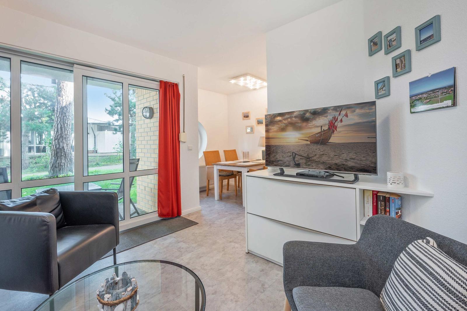 Wohnbereich mit TV, Sofa und Blick auf Garten. Rote Vorhänge und Essbereich im Hintergrund.