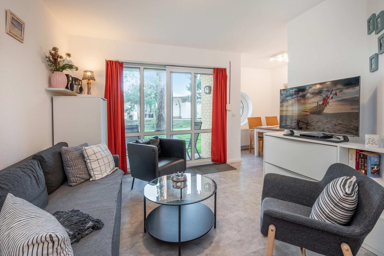 Wohnzimmer mit Sofa, Sesseln und TV. Große Fenster mit roten Vorhängen.