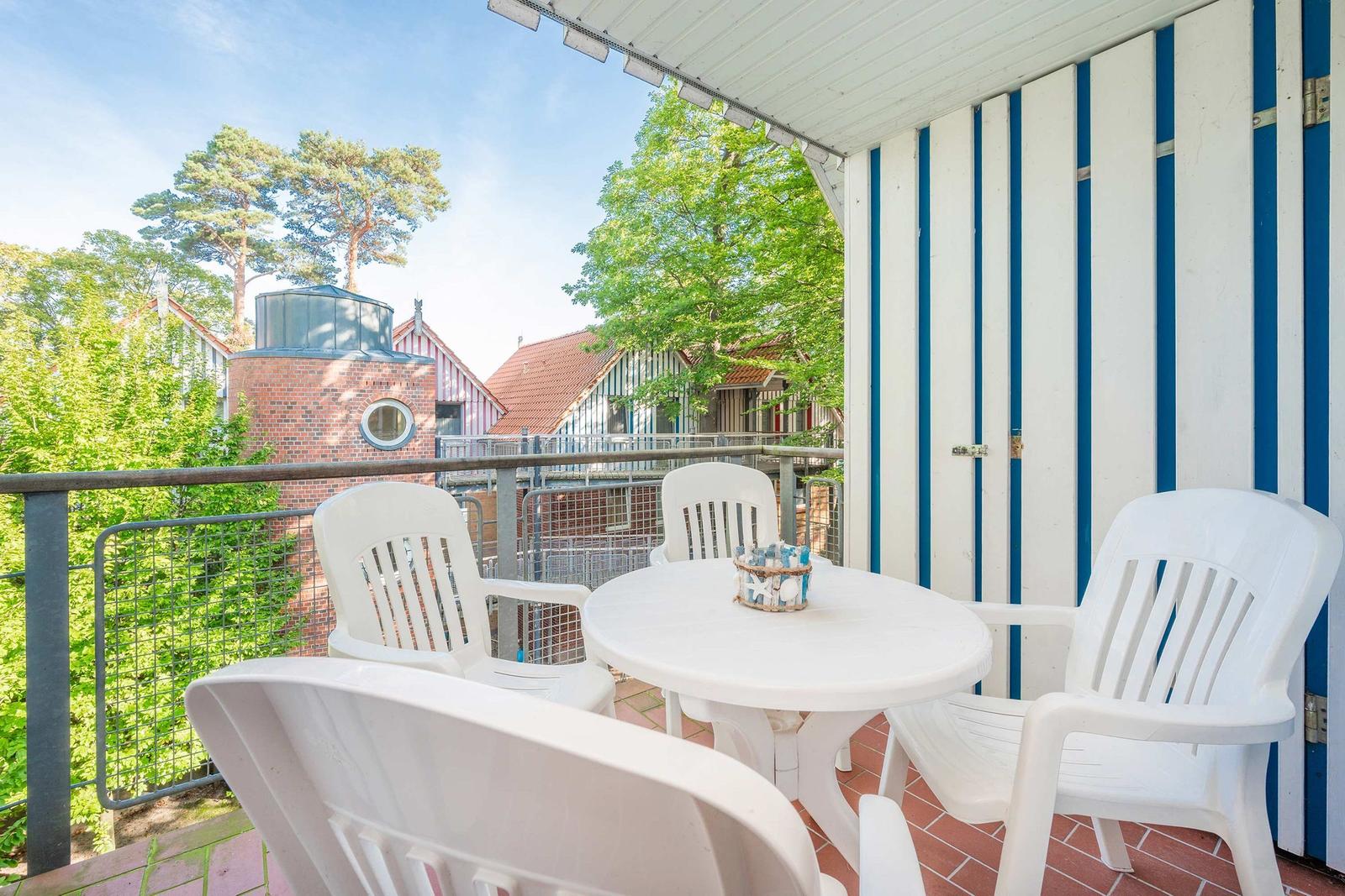 Terrasse mit weißem Tisch und Stühlen, blau-weiß gestreifter Wand, Blick auf Bäume und Gebäude.