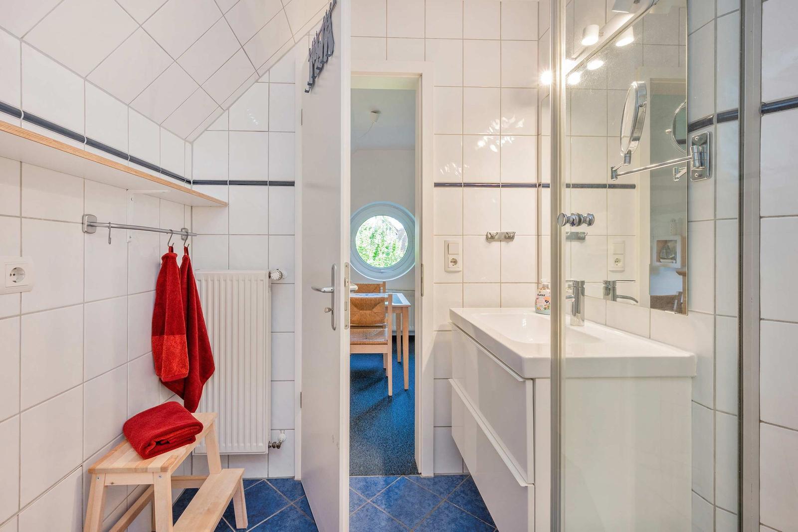 Badezimmer mit Dusche, Waschbecken und roten Handtüchern. Durchgang zu einem Raum mit runder Fensteröffnung.