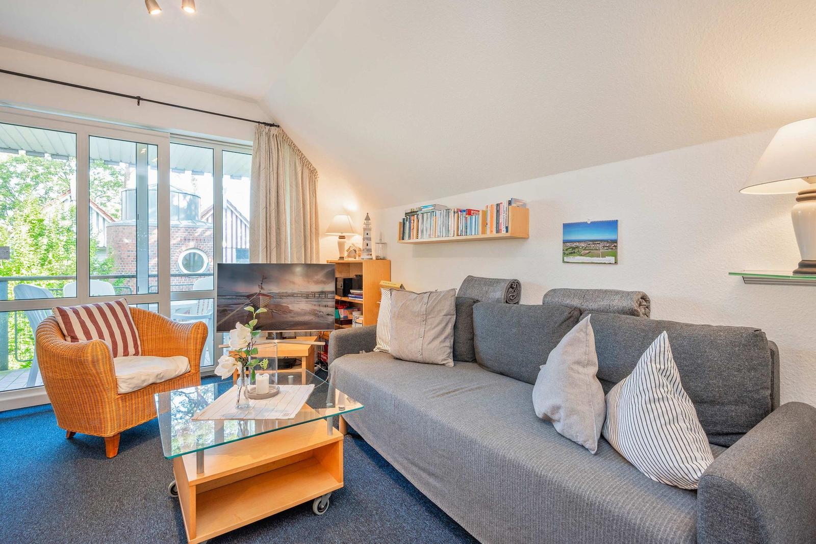 Wohnzimmer mit Sofa, Sessel, Glas-Tisch und Fernseher. Große Fenster mit Blick auf Balkon.