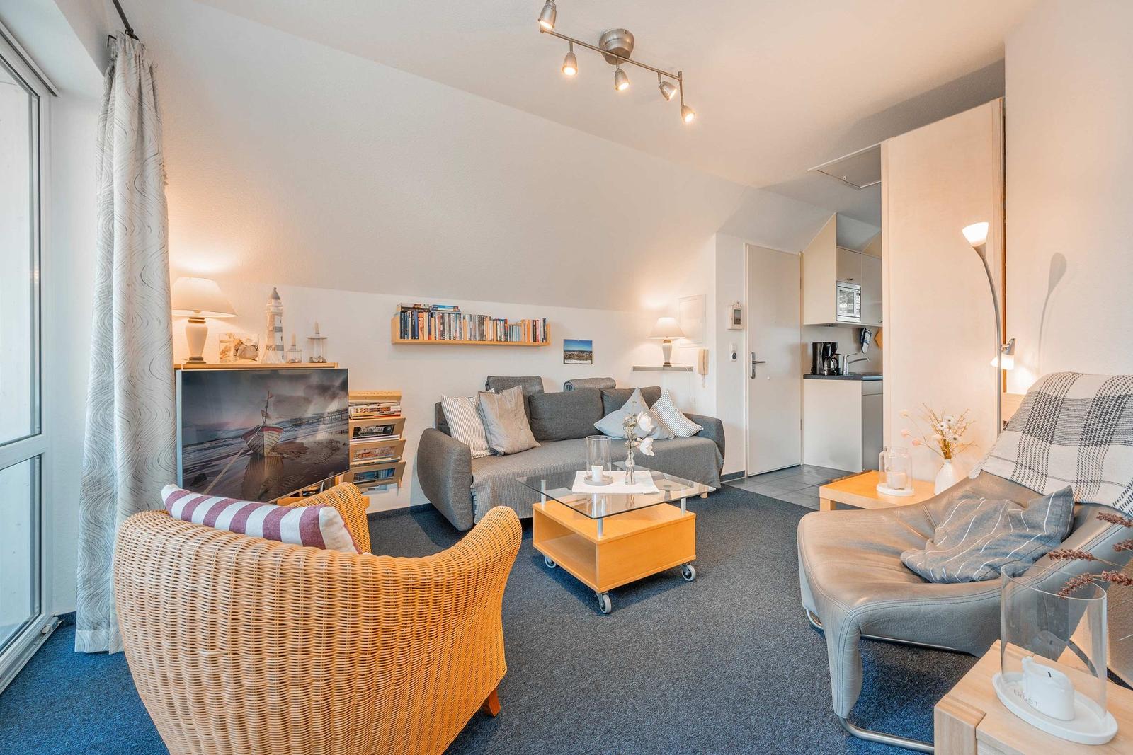 Wohnzimmer mit Sofa, Sessel und Glas-Tisch. Küche im Hintergrund. Fernseher und Bücherregal.