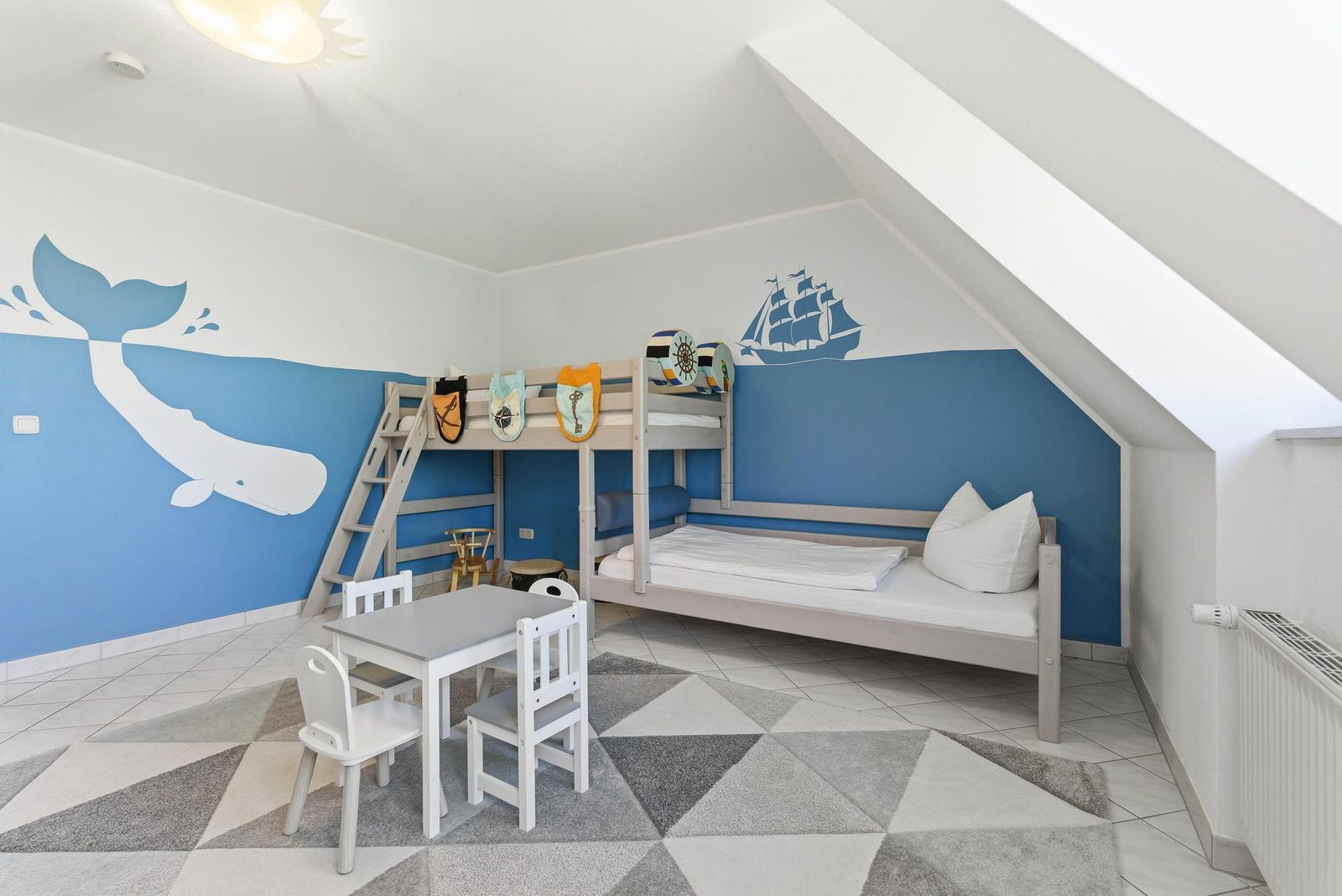 Kinderzimmer mit Doppelstockbett, Tisch, Stühlen und Wandmalereien von Wal und Schiff.