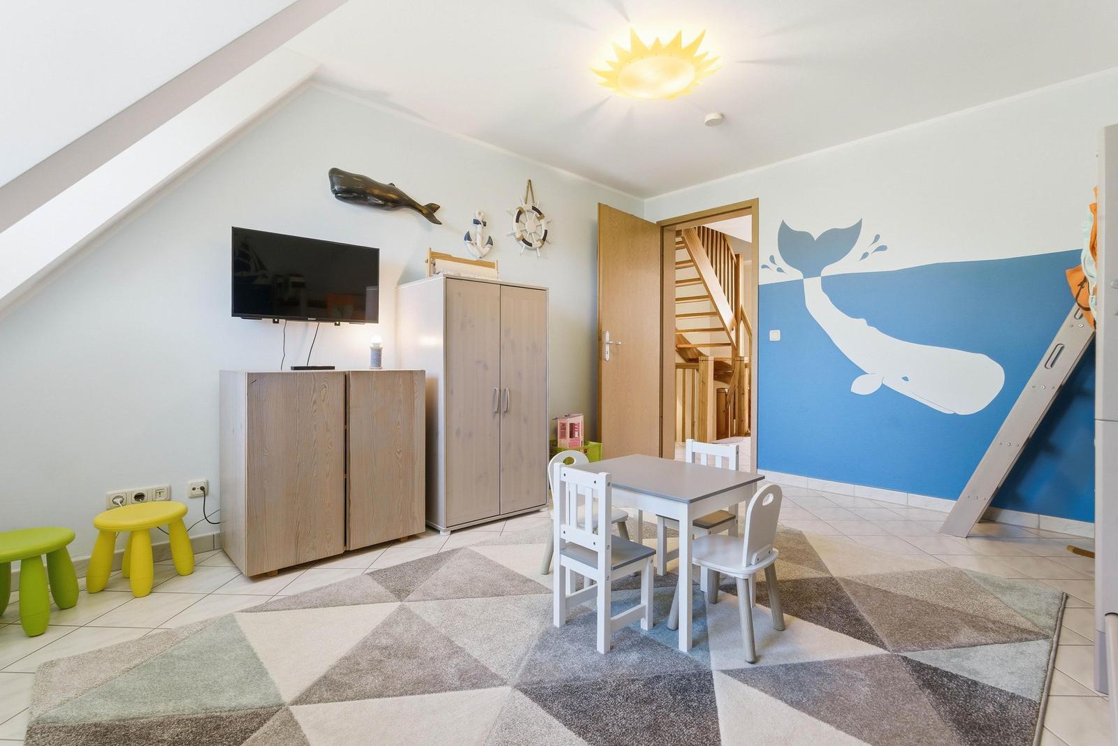 Kinderzimmer mit Wandmalerei, Tisch, Stühlen und TV. Holzregale und farbige Hocker.