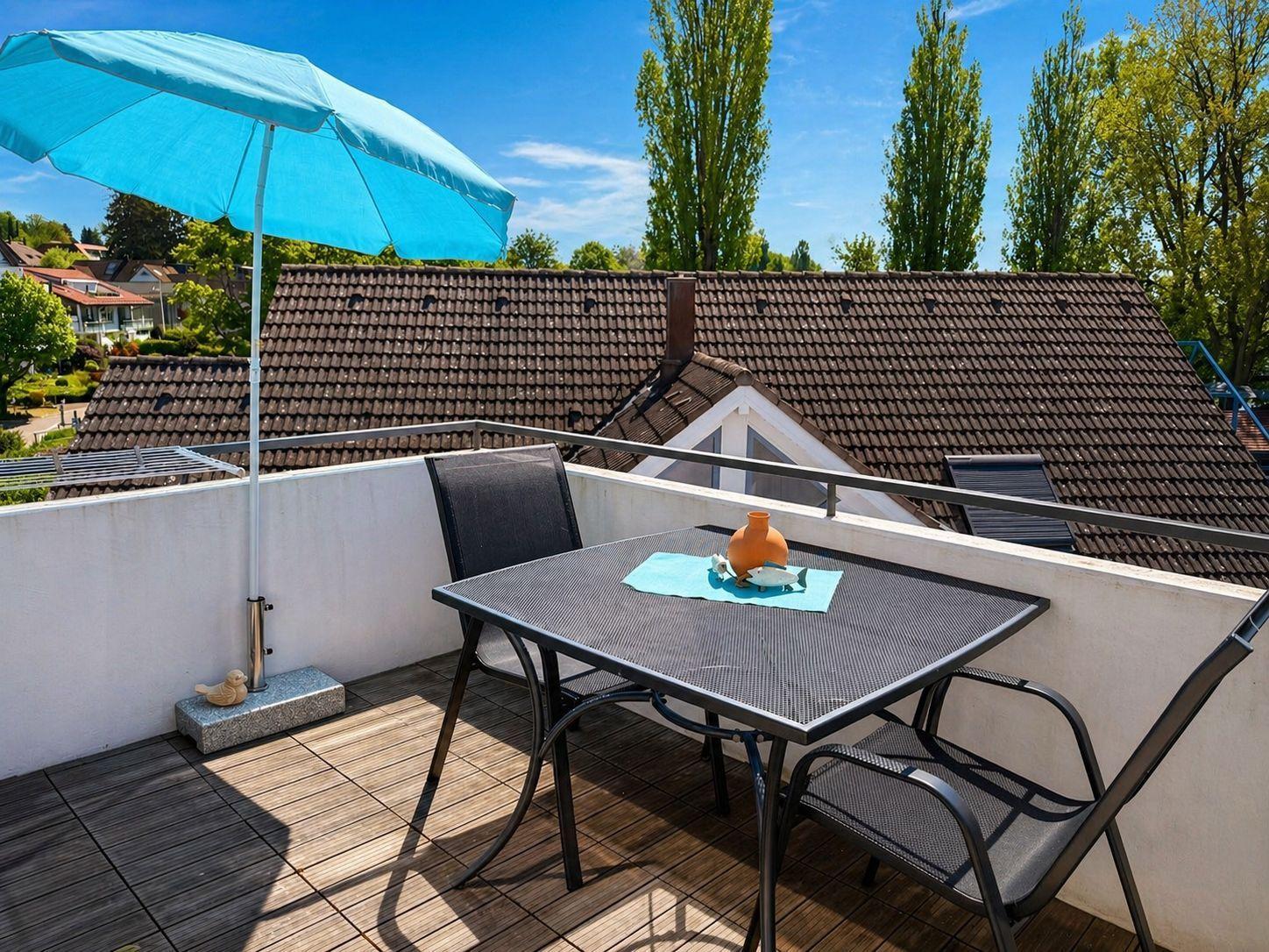 Terrasse mit Tisch, Stühlen und blauem Sonnenschirm. Blick auf Dächer und Bäume.
