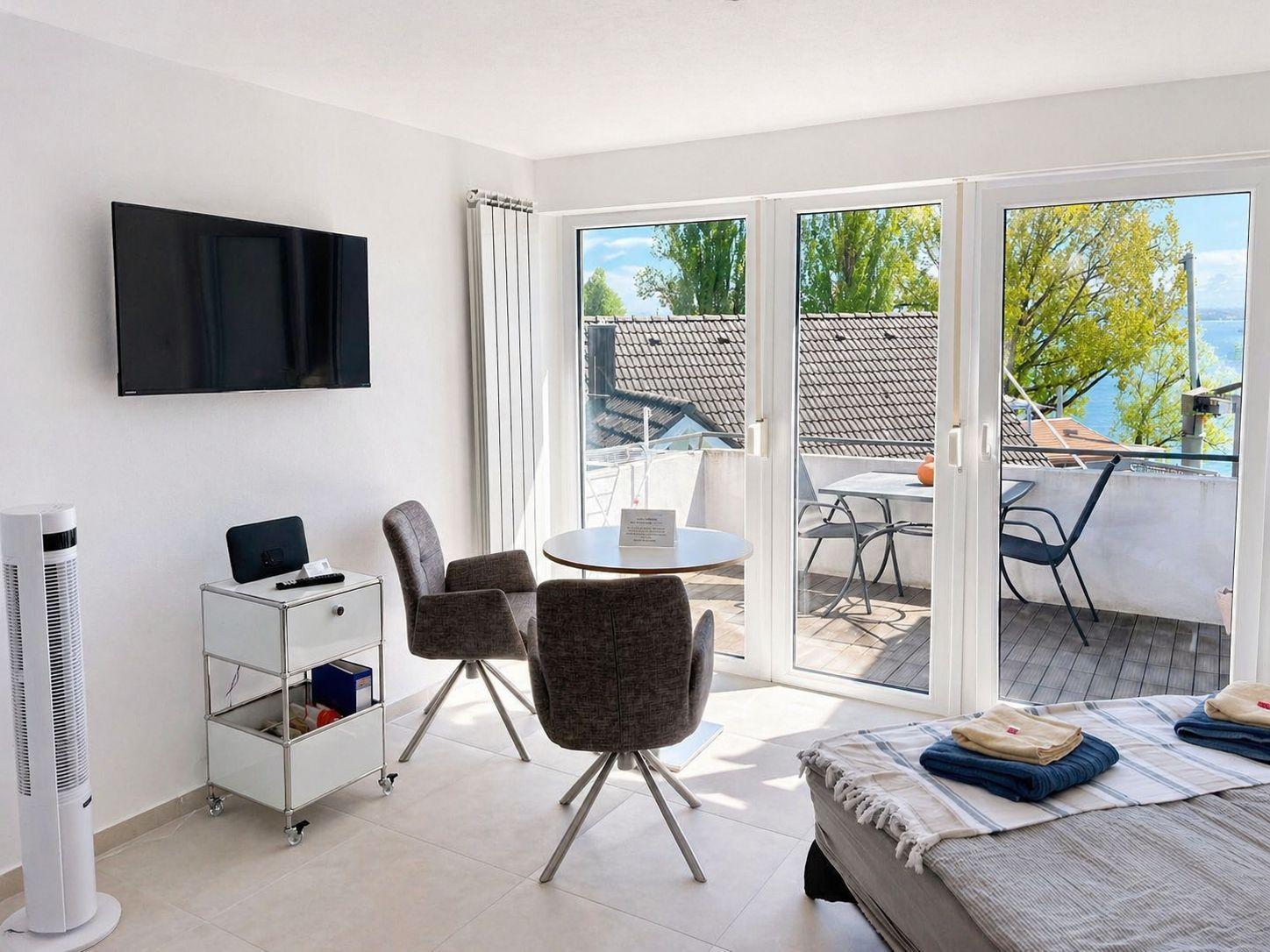 Zimmer mit TV, Tisch, Stühlen und Balkon mit Blick auf Wasser.