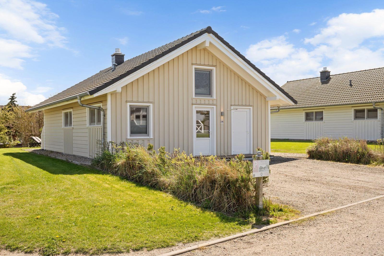 Ein hellfarbenes Haus mit weißer Tür und Fenstern, umgeben von Gras und einem Kiesweg.