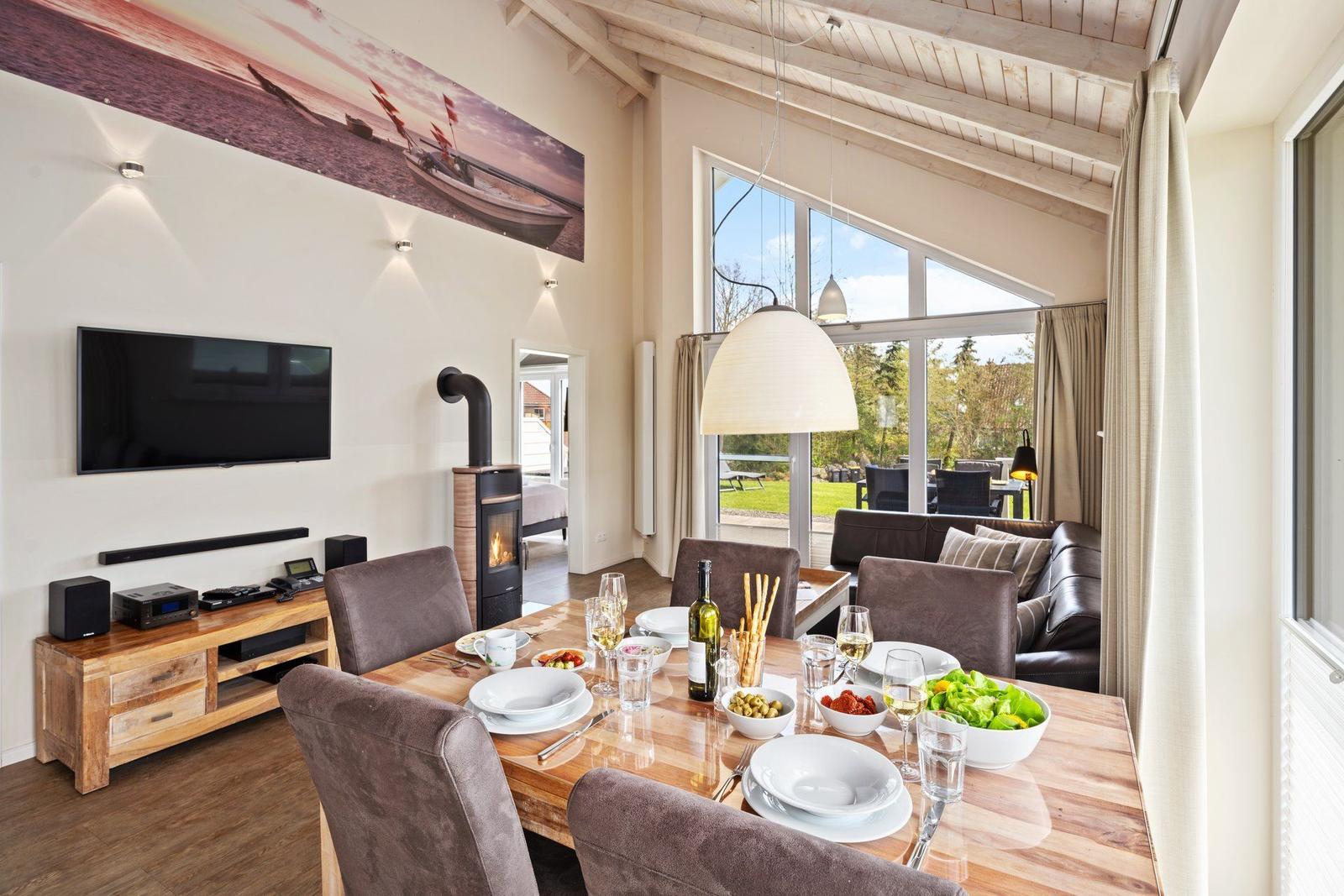 Esszimmer mit Holztisch, Sitzgelegenheiten und Blick auf Garten. Wandbild, TV und Kamin sichtbar.