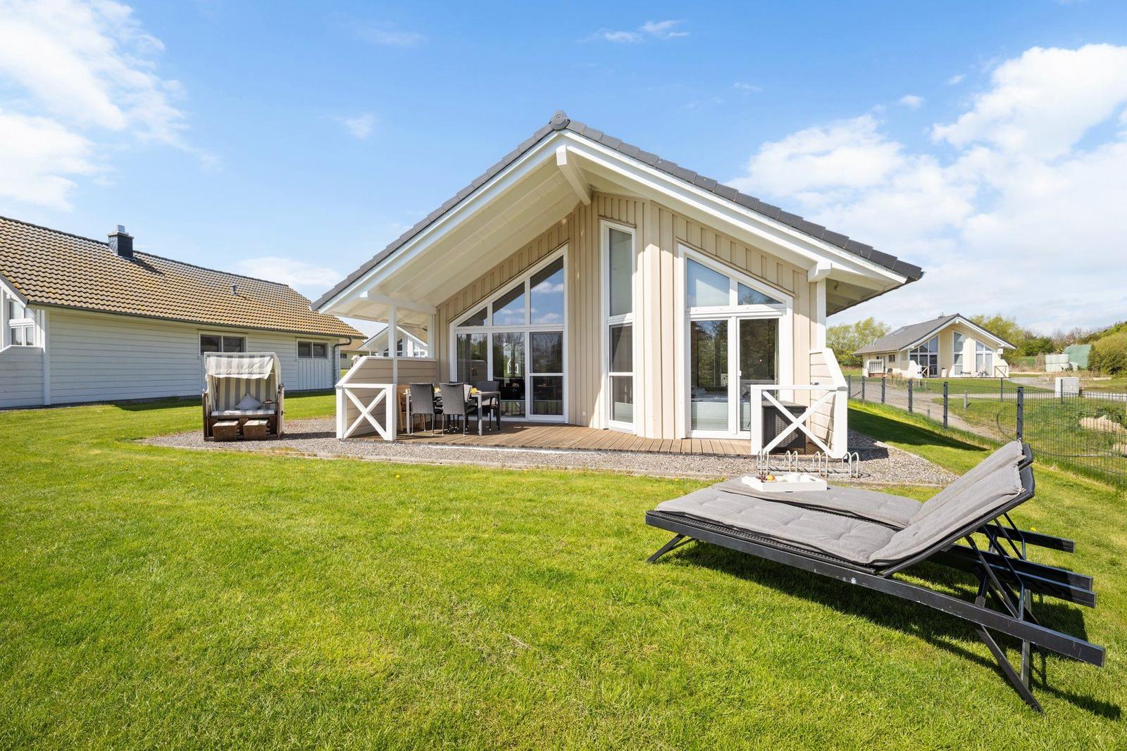Moderne Villa mit großem Garten, Terrasse und Liegestühlen. Glasfront öffnet sich zur Außenterrasse.