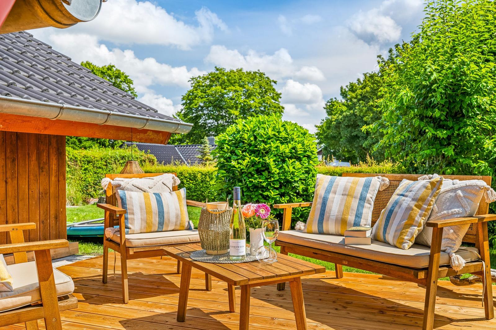 Holzterrasse mit Sitzgruppe, Tisch und Wein. Grüner Garten im Hintergrund.