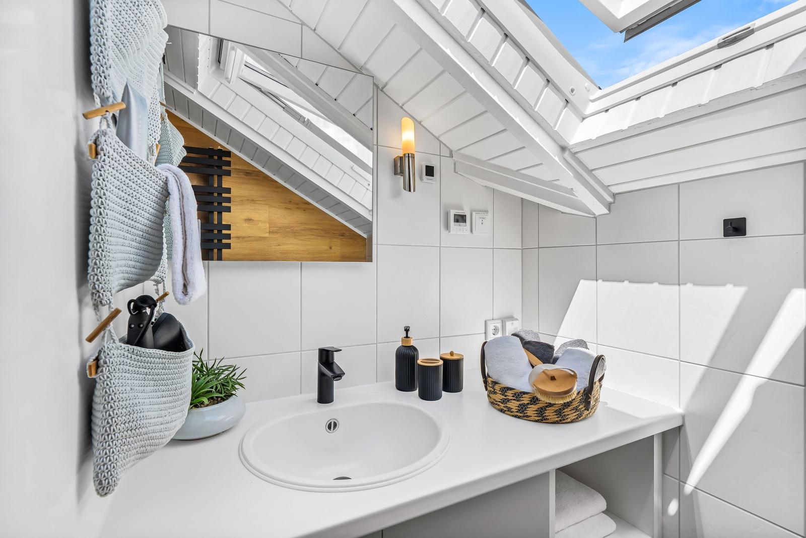 Badezimmer mit Waschbecken, Spiegel und Dachfenster. Weiße Fliesen und moderne Accessoires.