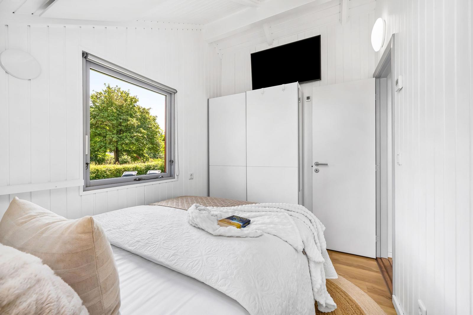 Schlafzimmer mit weißer Einrichtung, Bett, Kleiderschrank und Fenster mit Blick auf Bäume.