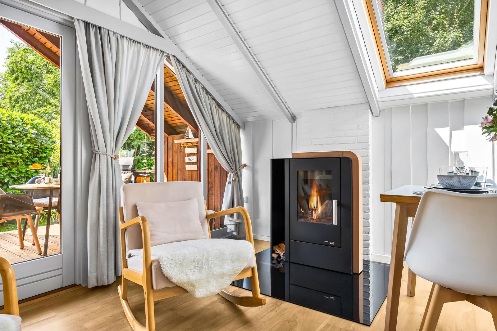 Ein Raum mit Sessel, Kamin und Tisch. Durch Fenster ist ein Garten sichtbar.