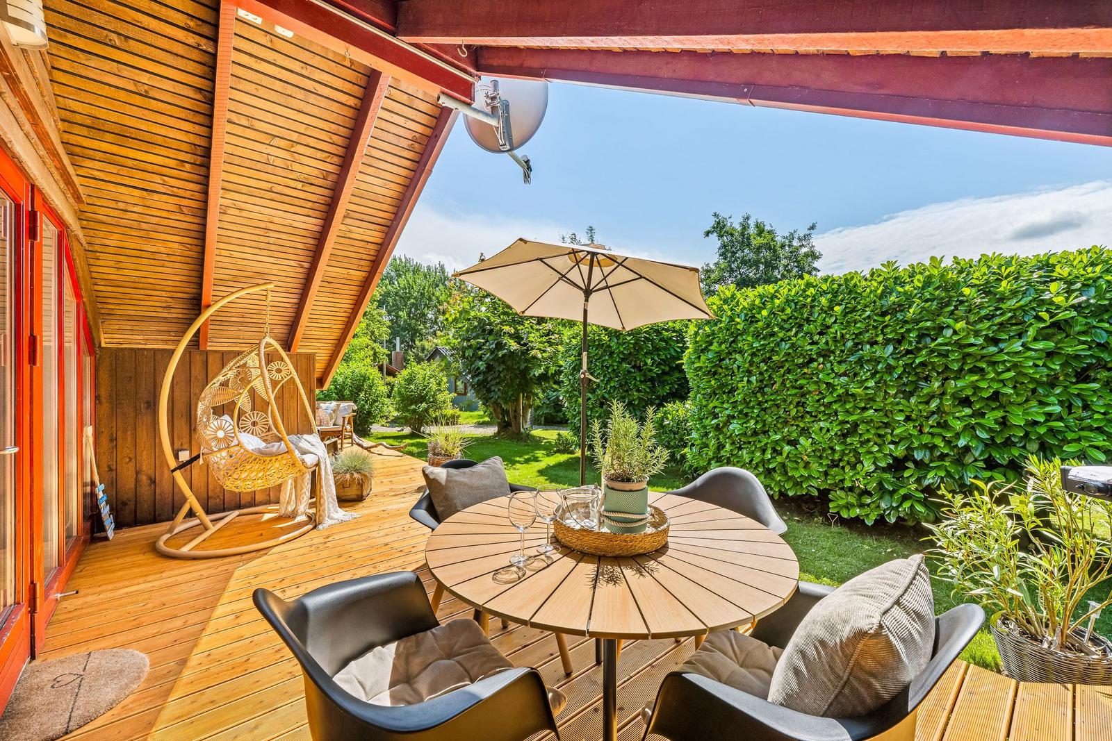 Terrasse mit Tisch, Stühlen, Sonnenschirm und Hängesessel unter Holzüberdachung.
